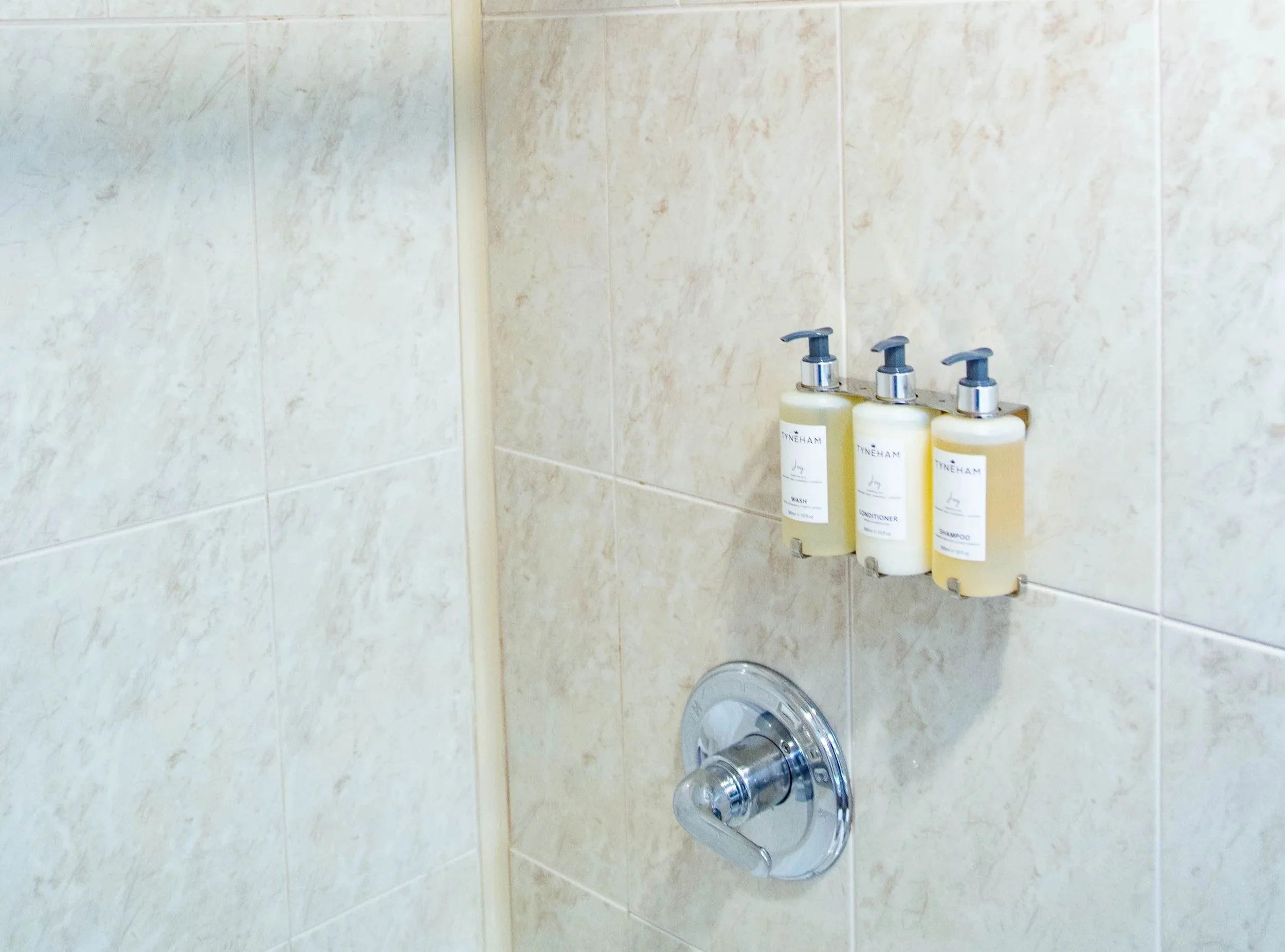 Close-up of a beige tiled shower wall with three bottles of toiletry products labeled shampoo, conditioner, and body wash, and a silver shower handle below.