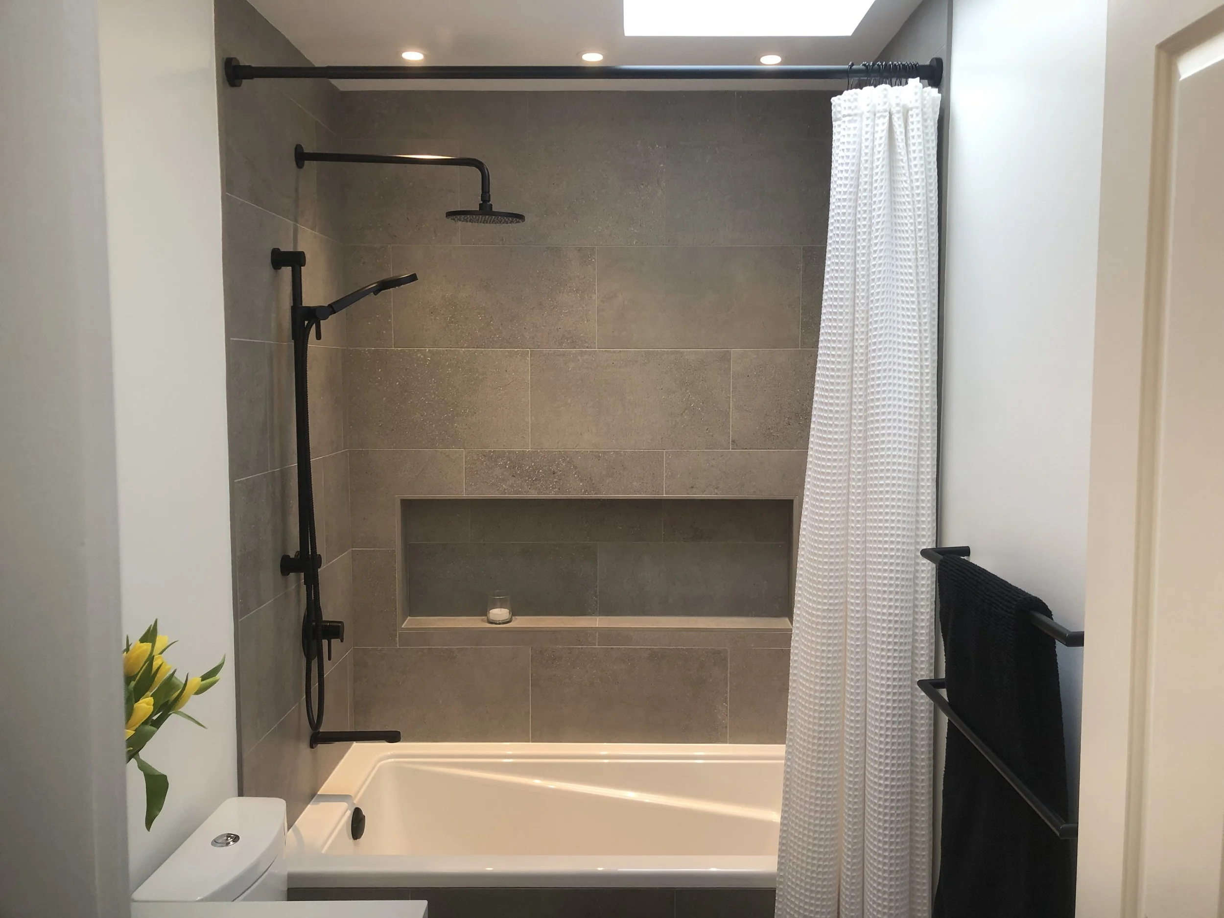 A modern walk-in shower with a black showerhead and handle, beige tiled walls, a white shower curtain with a textured pattern, built-in niche with a candle, a black towel rack, and a small bouquet of yellow flowers on a ledge.