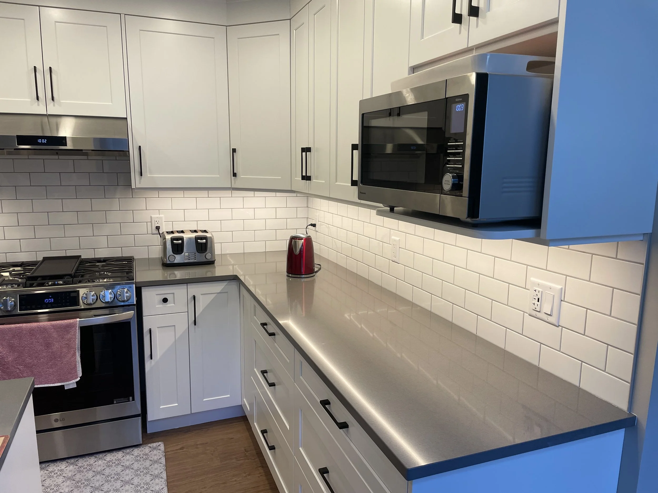 Modern kitchen with white cabinets, stainless steel appliances including microwave and stove, white subway tile backsplash, and a red kettle on the gray countertop.