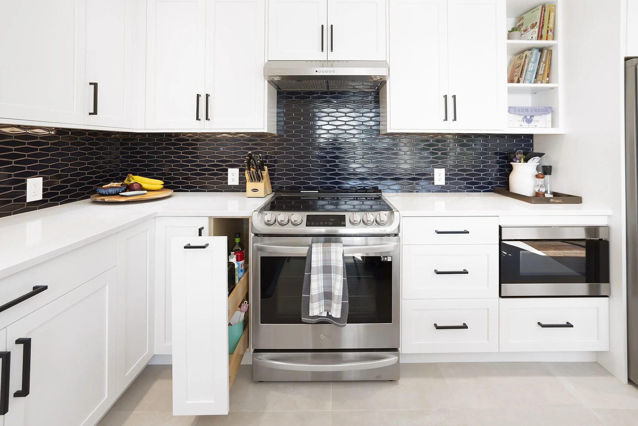 A modern kitchen with white cabinets, black hardware, and a black hexagonal tile backsplash. Stainless steel stove with a gray checkered towel hanging from the handle. On the left, a counter with bananas, a watermelon, and an onion. On the right, a w