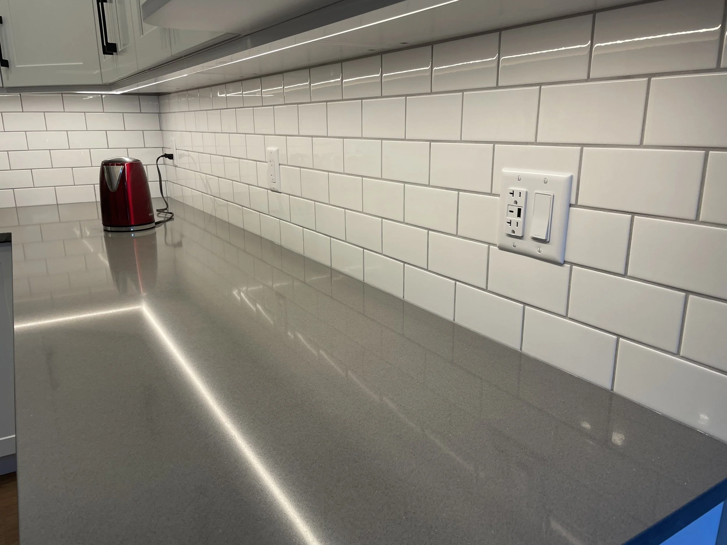 Kitchen countertop with a red electric kettle, white tiled backsplash, wall outlets, and a reflective gray surface.