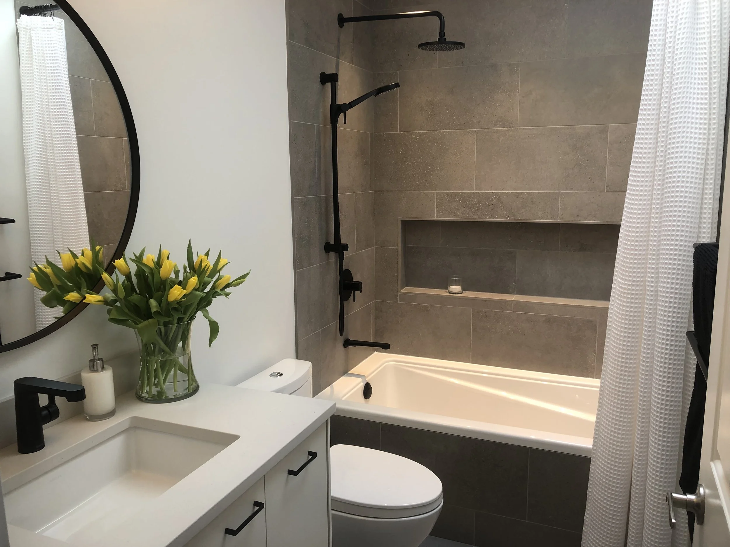 A modern bathroom with a white vanity that has a black faucet, a spray of yellow tulips in a glass vase, and a soap dispenser. There is a large oval mirror above the vanity. The background features a shower area with gray tiles, a built-in shelf, and a black showerhead and handle. White curtains frame the sides of the image.