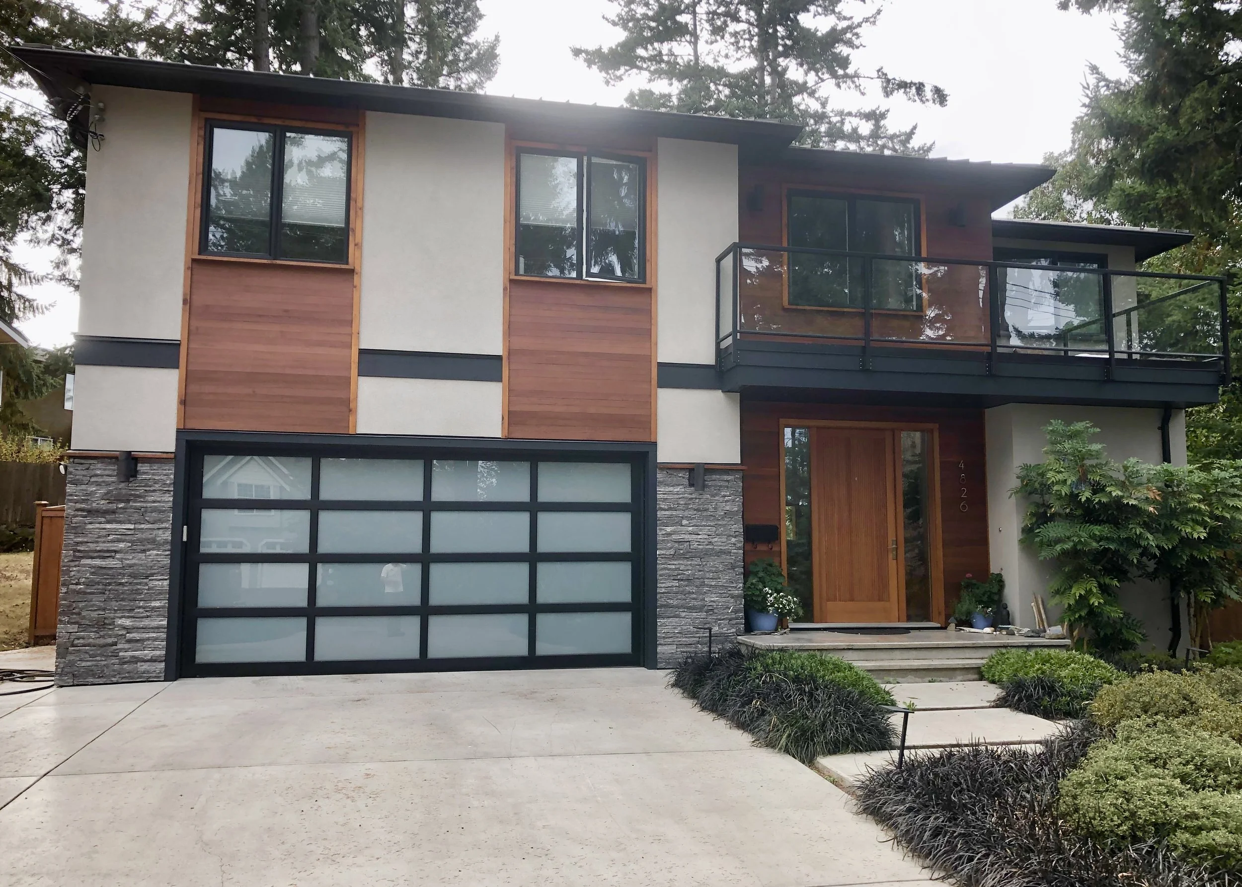 A modern two-story house with a combination of wood, glass, and stone exterior, featuring large windows, a glass balcony, and a garage with a dark frame and frosted glass door, surrounded by greenery and trees.