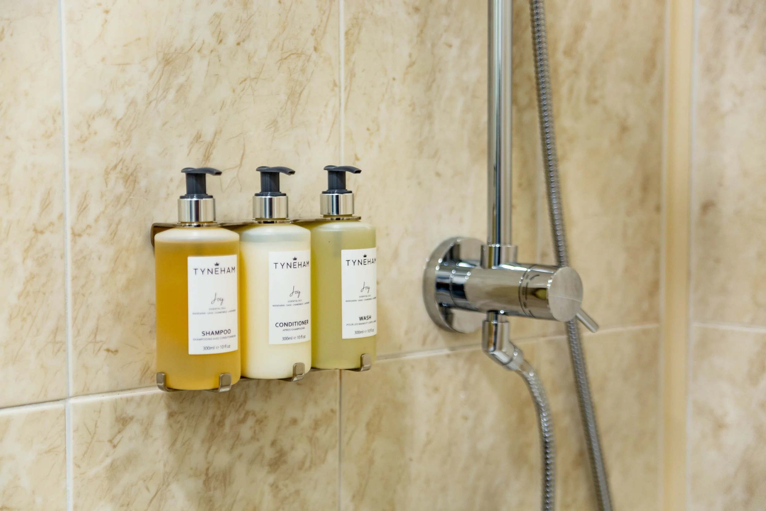 Soap and shampoo bottles labeled 'Tyneham Joy' on a shower wall next to a chrome shower faucet and hose.