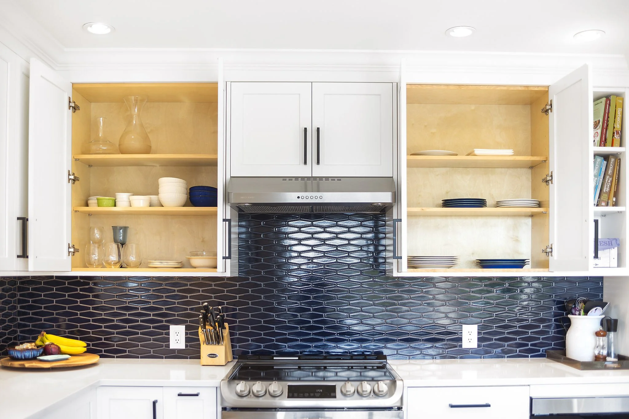 White kitchen cabinets with open shelves, a stainless steel range hood, a stove, and a black hexagonal tile backsplash. Countertop with fruit, a knife block, and kitchen utensils.