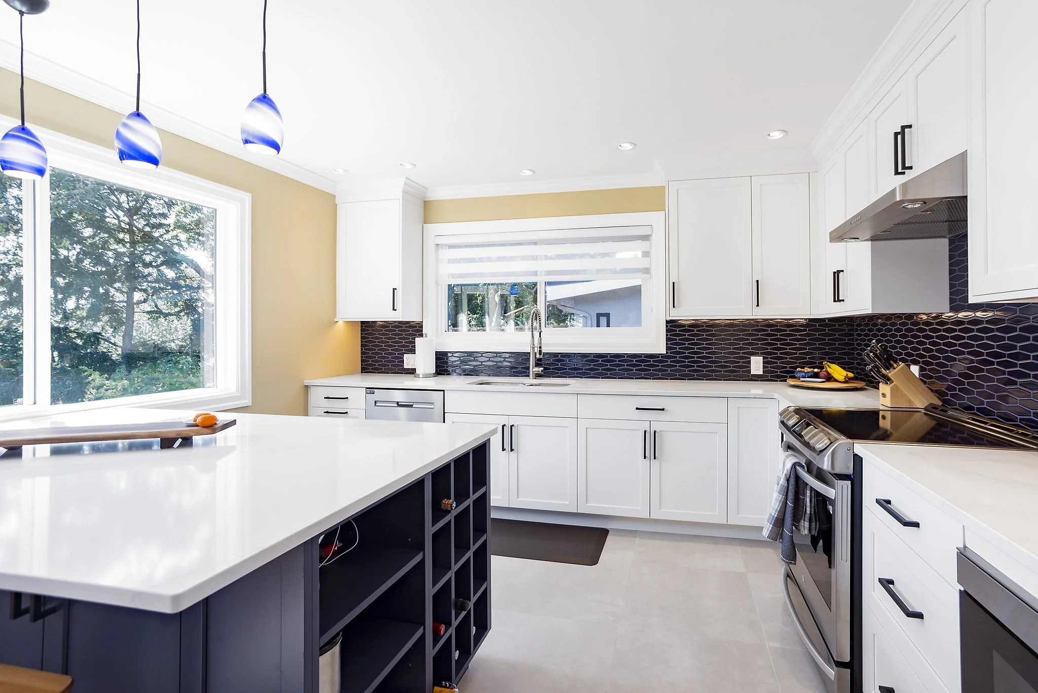 Modern kitchen with white cabinets, black backsplash, large windows, and a white island with a dark lower section. Blue pendant lights are hanging from the ceiling.