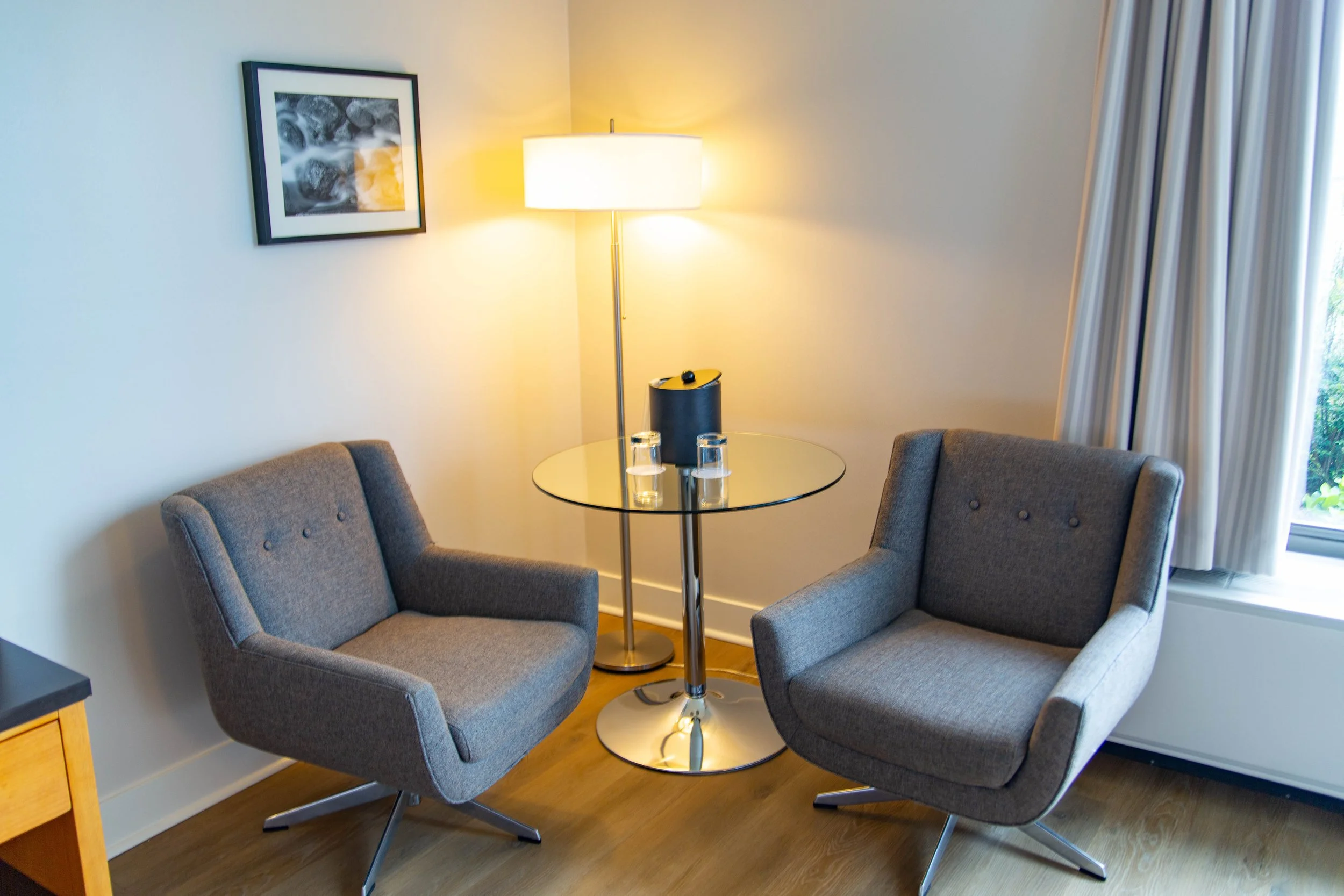 Two gray upholstered armchairs around a small round glass table with a lamp and two glasses inside a room with a framed photograph on the wall and a window with curtains.