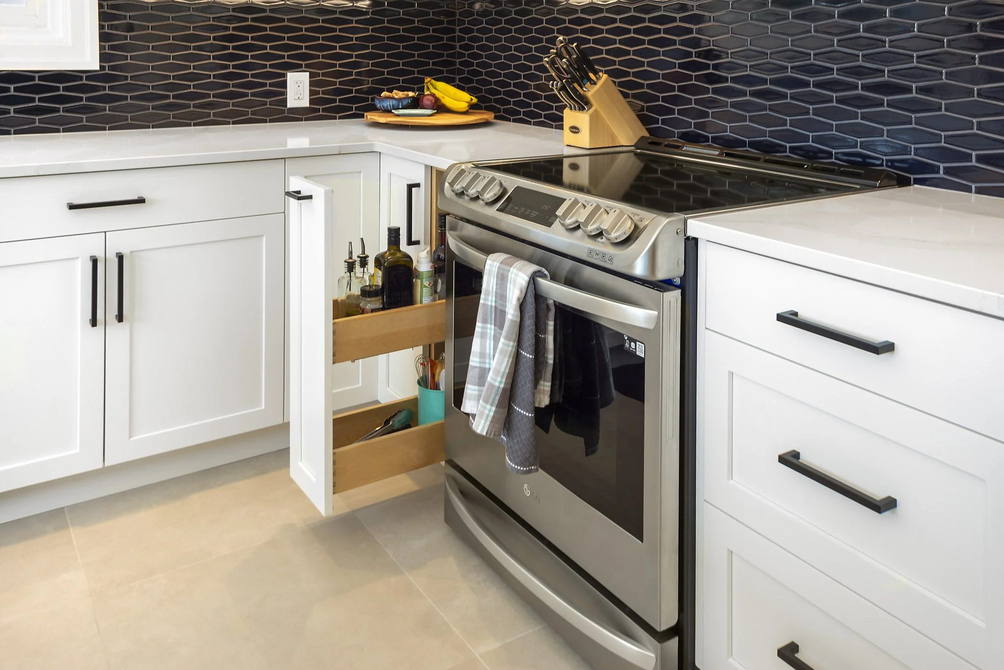 Modern kitchen with white cabinets, black hexagonal tile backsplash, and stainless steel oven. Open cabinet drawer with kitchen utensils and bottles, countertop with fruit and decorative items, and a set of knives on a wooden block.