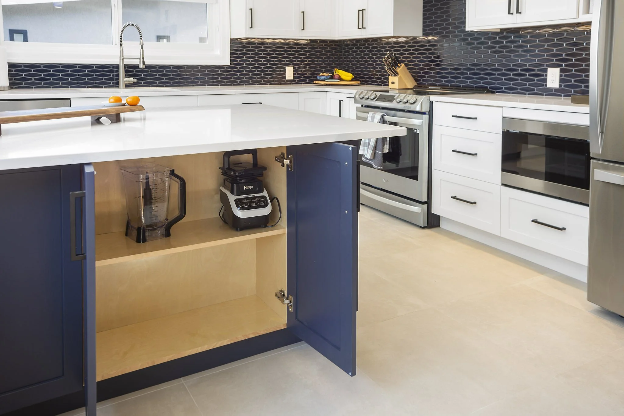 Modern kitchen with white cabinets, black split tile backsplash, and stainless steel appliances. An island features a dark blue cabinet with an open shelf containing a blender and a box. There is a wooden tray with two oranges on the island. The coun
