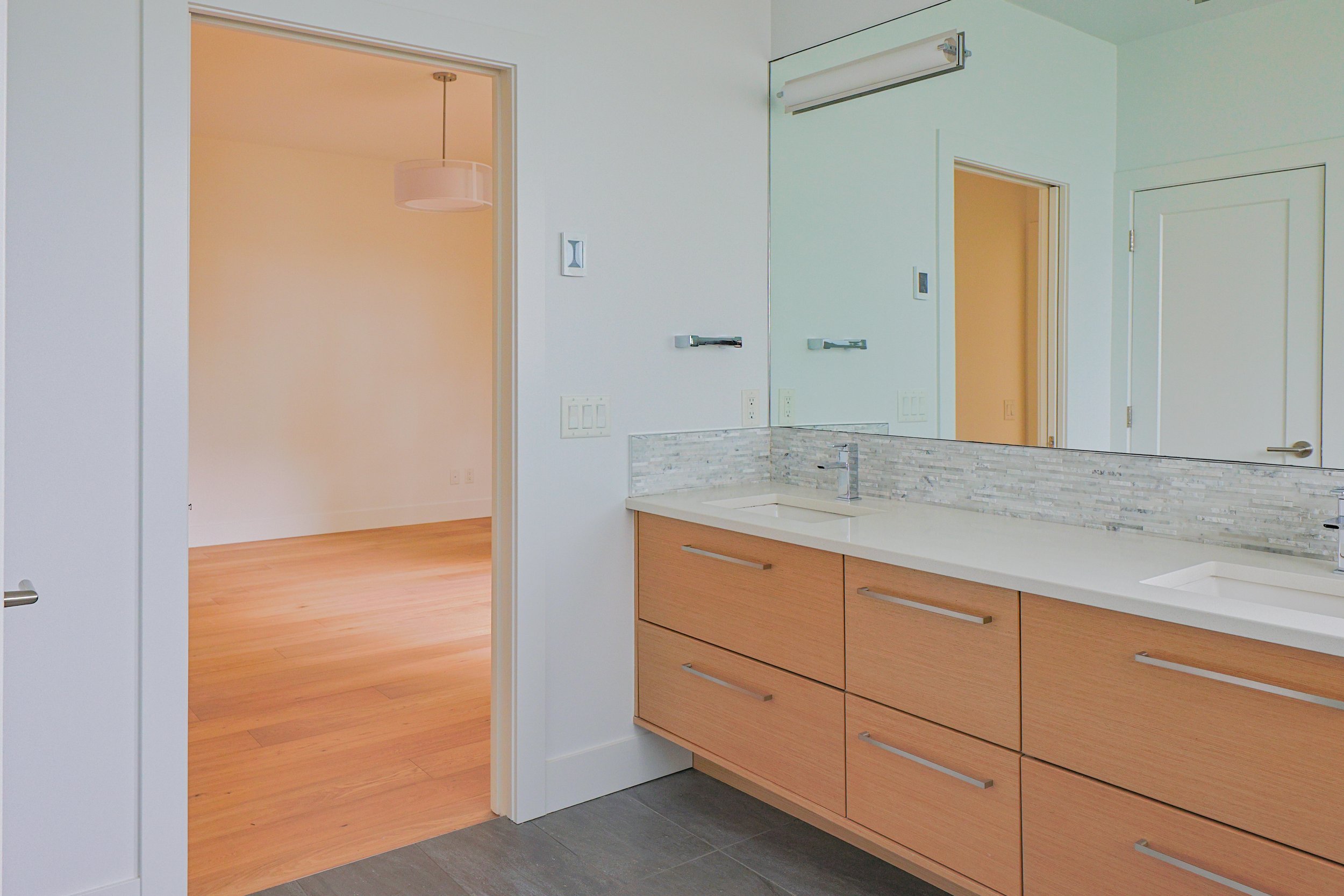 Modern bathroom with a large mirror, wooden vanity with drawers, white countertop, and mosaic tile backsplash. Open doorway leads to a room with wood flooring and a hanging light fixture.