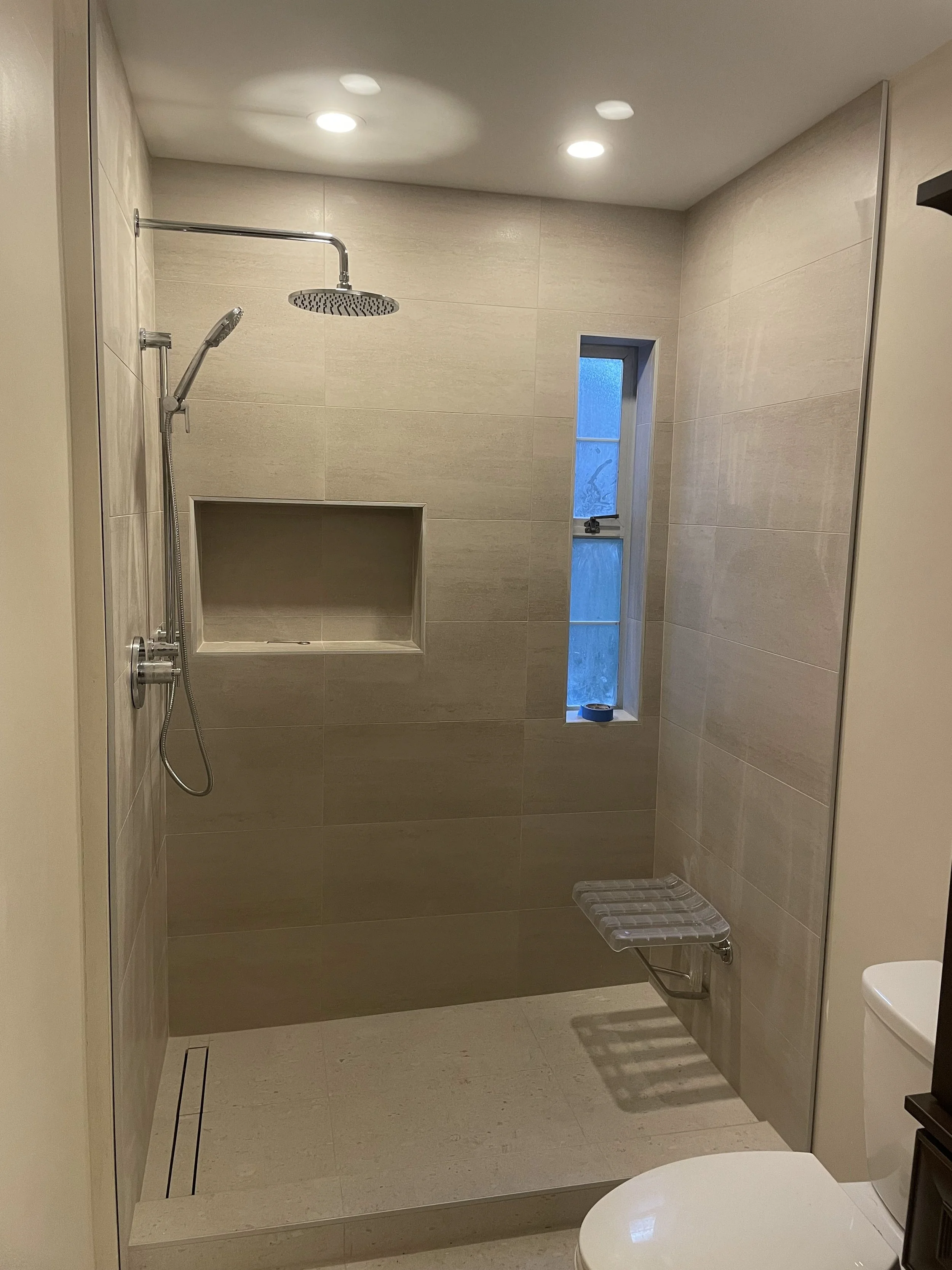 Modern bathroom with beige tiled walls and floor, a walk-in shower with a rain showerhead, small window, and a transparent shower shelf.