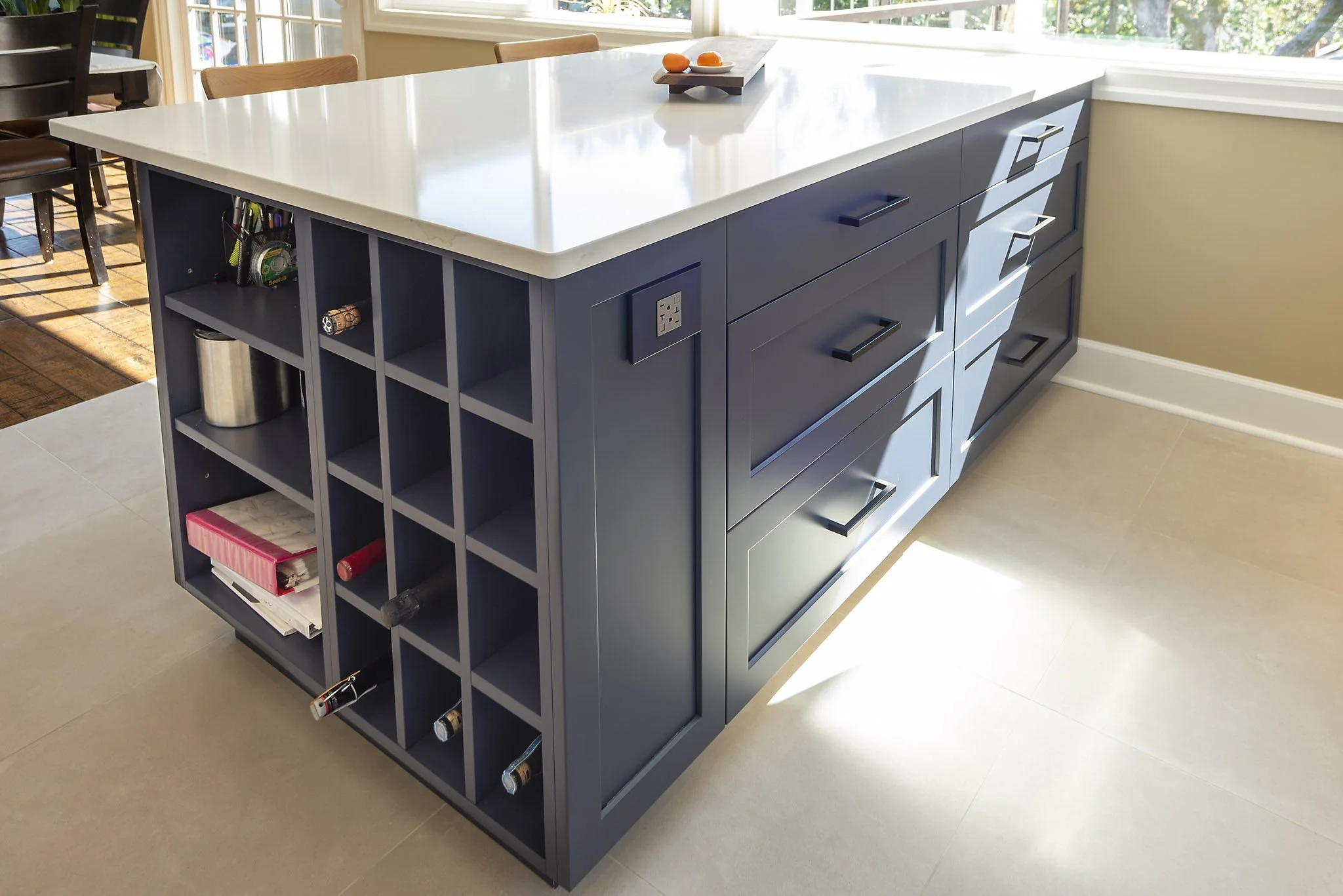 Kitchen island with a white countertop and blue cabinets, open shelving on one side with various items including pens, books, and cans, and a power outlet on the side, with sunlight streaming through a window.