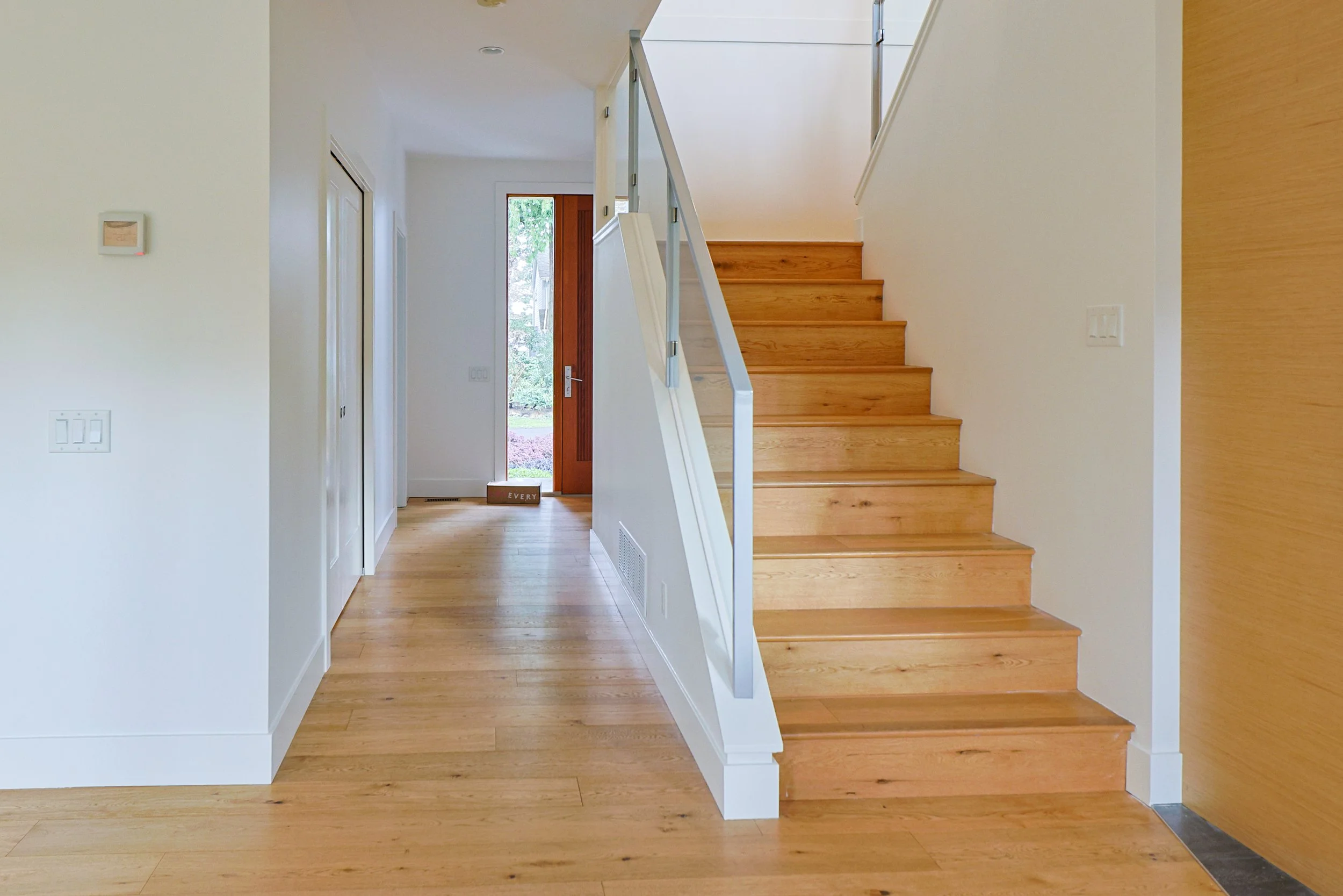 Interior view of a modern home with a staircase, wooden flooring, and a glass-paneled front door leading outside. The walls are painted white.