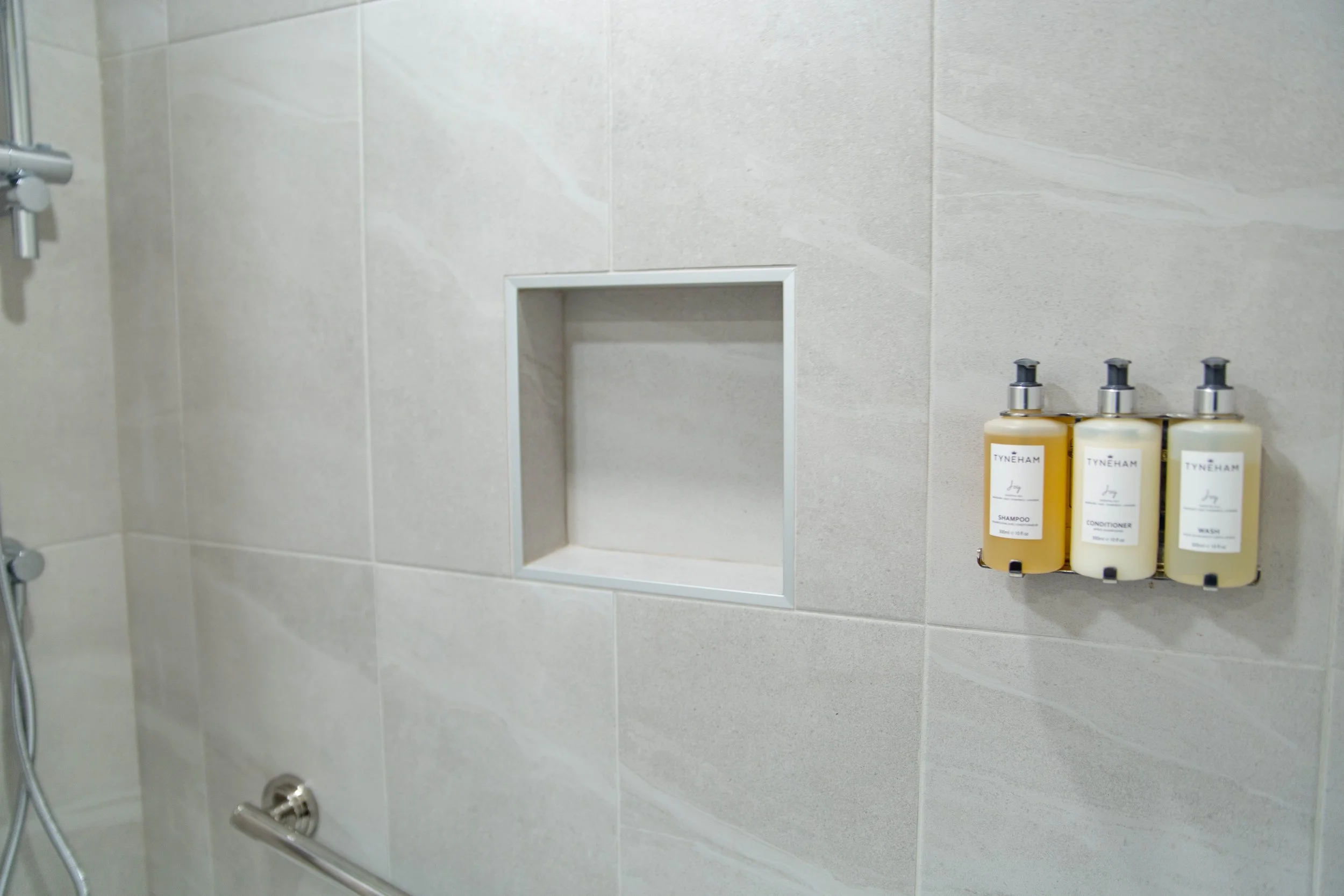 Close-up of a tiled bathroom shower wall with a built-in shower shelf holding three bottles of Tyneham shampoo, conditioner, and wash.