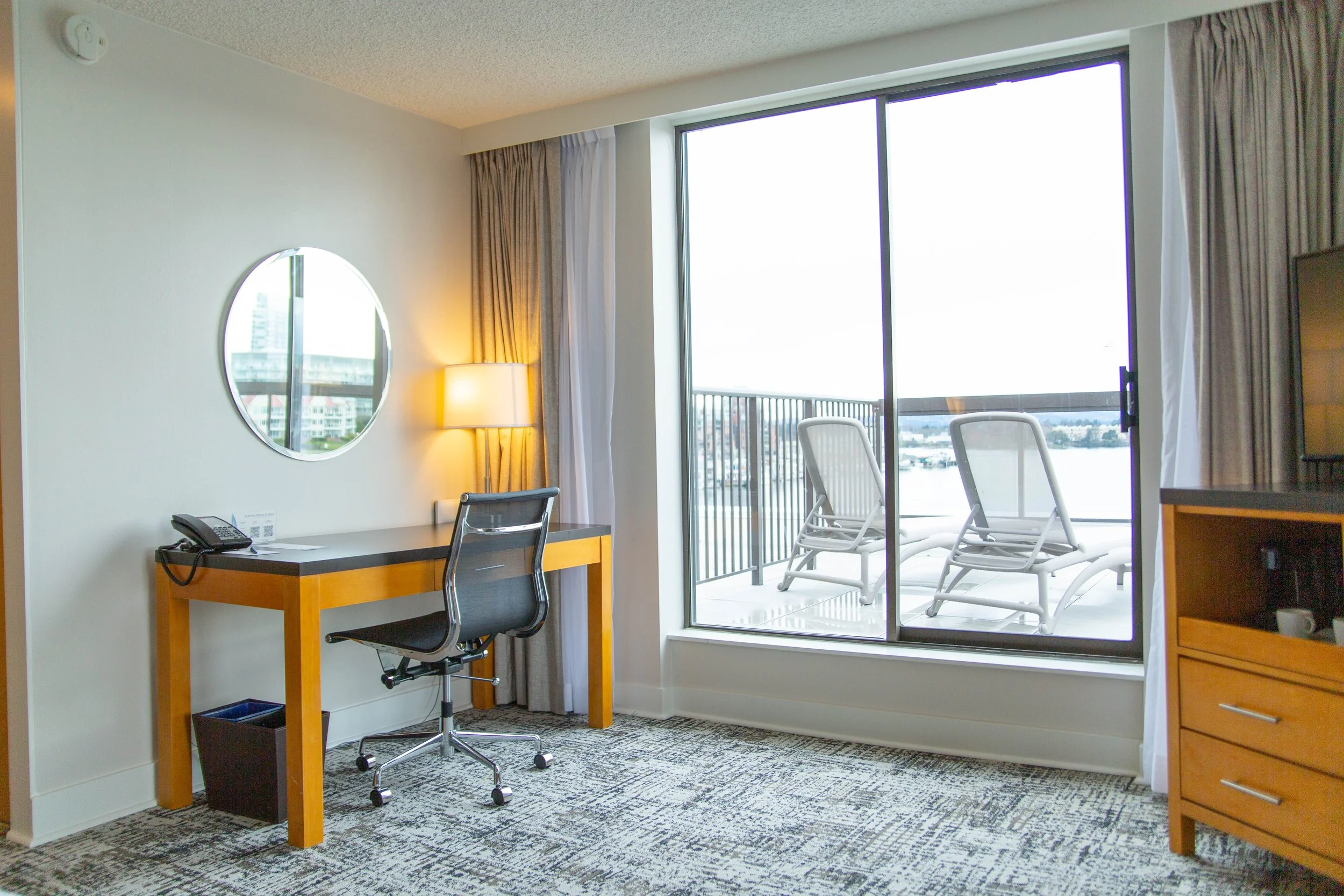 hotel room workspace with a desk, office chair, lamp, and balcony with two lounge chairs overlooking a river
