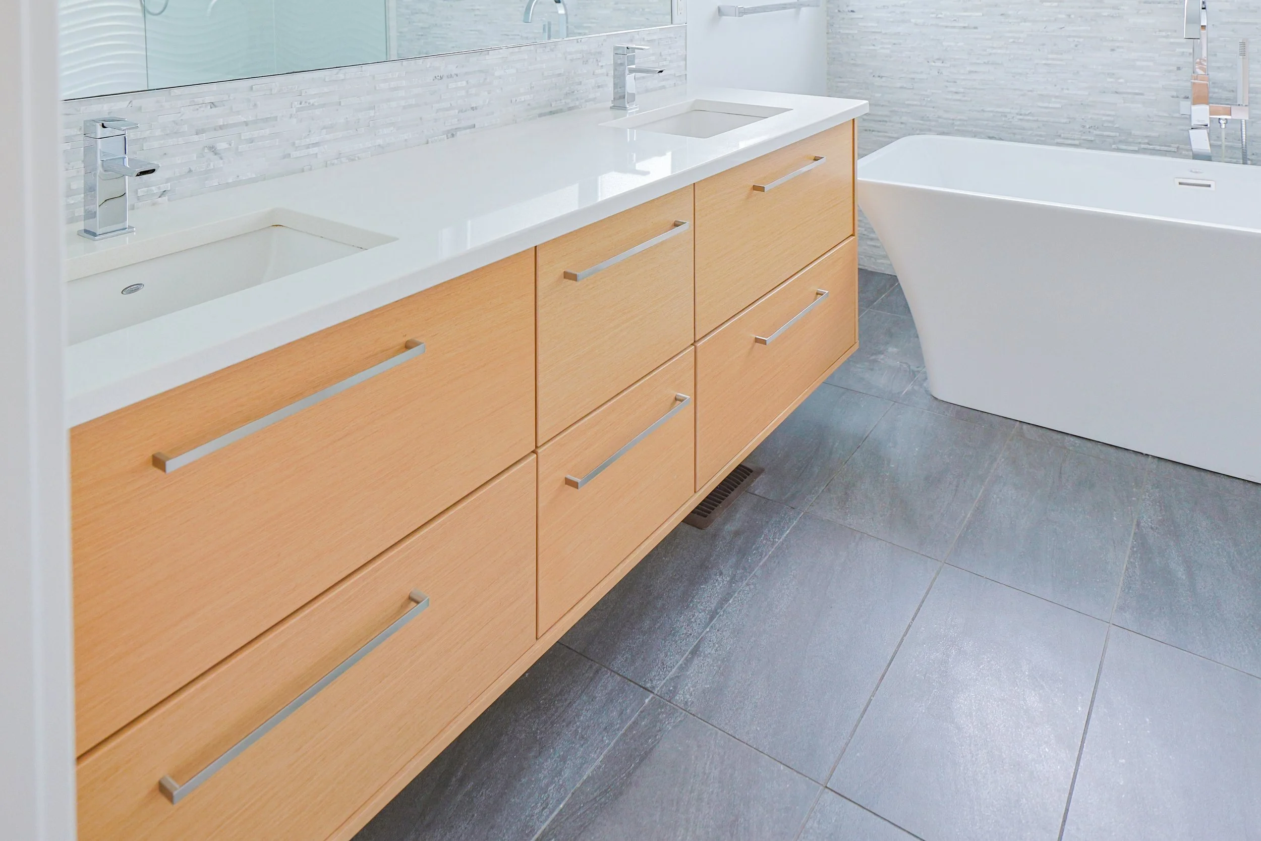 Modern bathroom with a double vanity with light wood cabinets, white countertops, and silver handles. Two rectangular sinks with silver faucets. Large mirror above the vanity. To the right, a white freestanding bathtub with a silver faucet and a buil