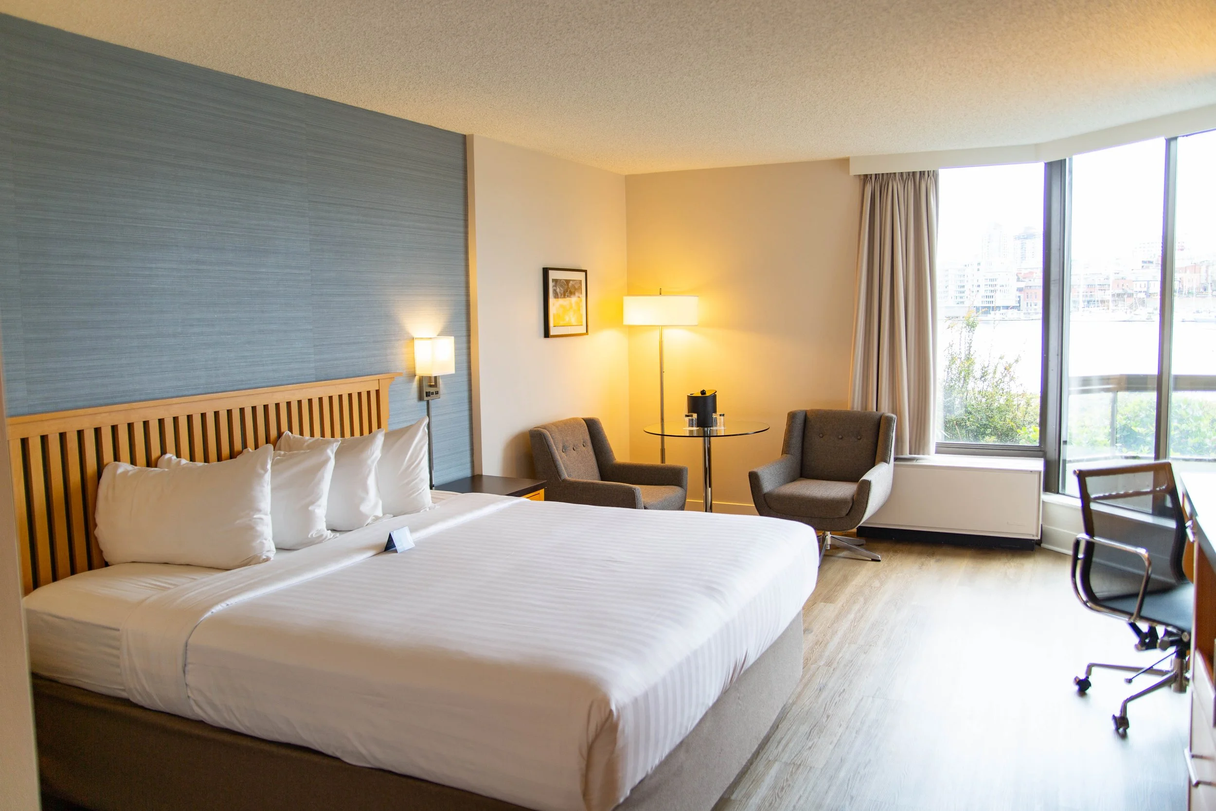 Interior view of a hotel room featuring a large bed with white linens, a wooden headboard, two bedside lamps, a small dining table with two chairs, and two gray armchairs near a large window with curtains. City buildings can be seen outside the windo
