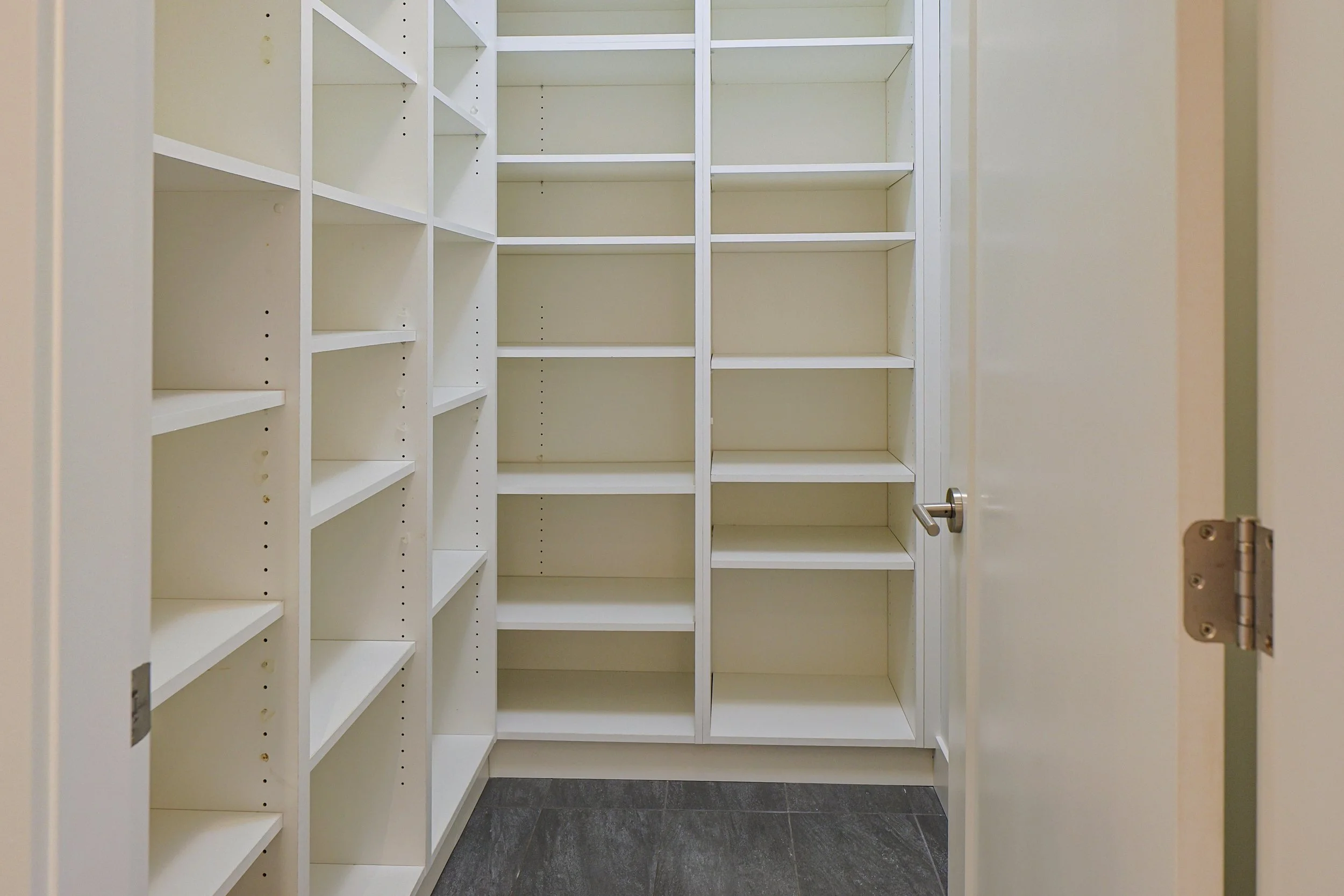Empty white built-in closet with multiple shelves, open door with silver handle, gray tile floor, beige walls.