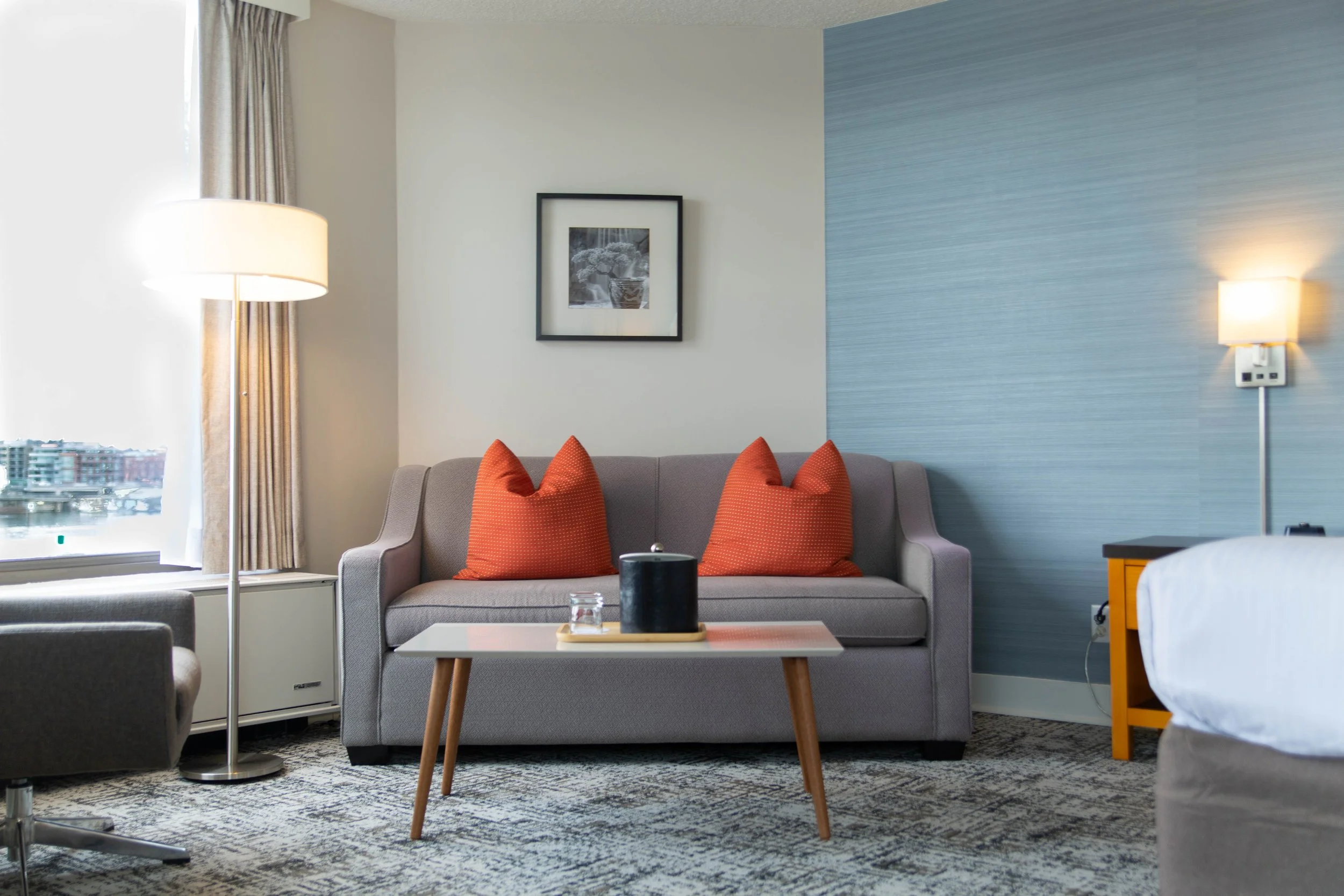 Modern hotel living room with a beige sofa decorated with red pillows, a wood coffee table, a framed black-and-white photograph on the white wall, a window with beige curtains, a table lamp, and a textured blue accent wall.