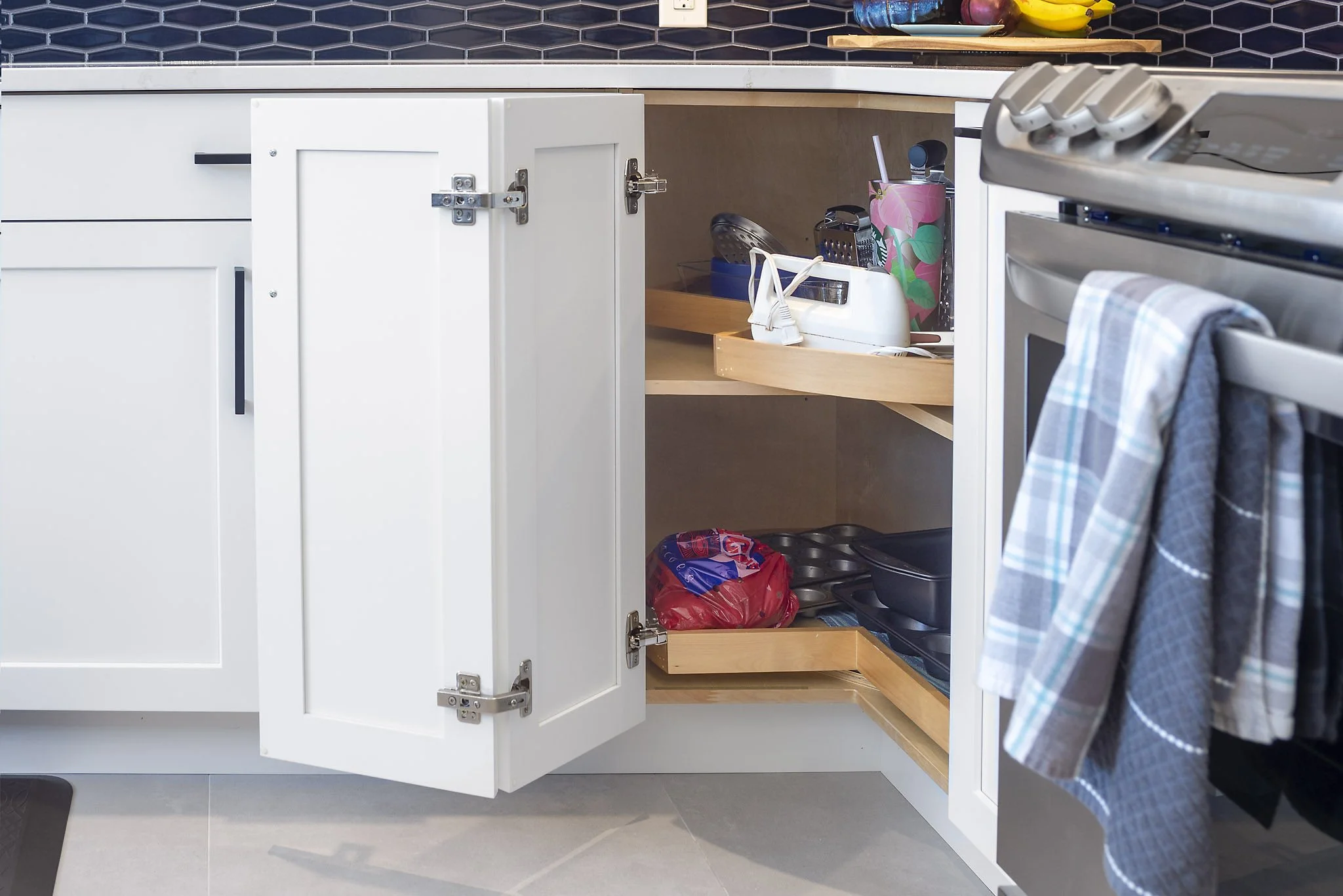 Kitchen cabinet with open door revealing pantry and various household items such as a bag of potato chips, kitchen tools, a toaster, and dish towels hanging on the oven handle.