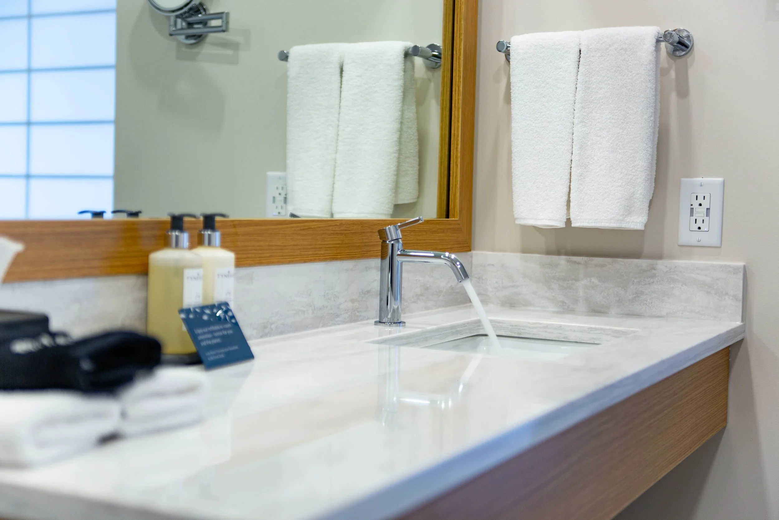 Bathroom sink with a chrome faucet, two white towels hanging on a towel rack, bottles of toiletries on the counter, a mirror, an electrical outlet, and a soap dispenser in a hotel or home bathroom.