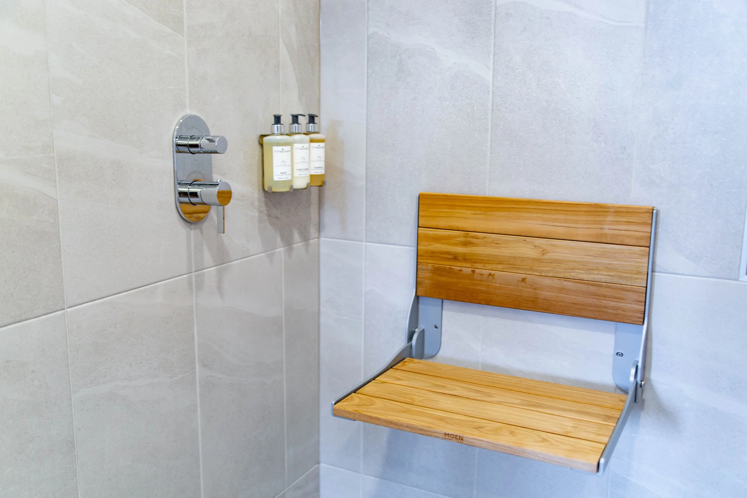 A bathroom corner with three Bvlgari toiletries bottles on a wall-mounted holder, a wooden bench with metal legs, and tiled walls.