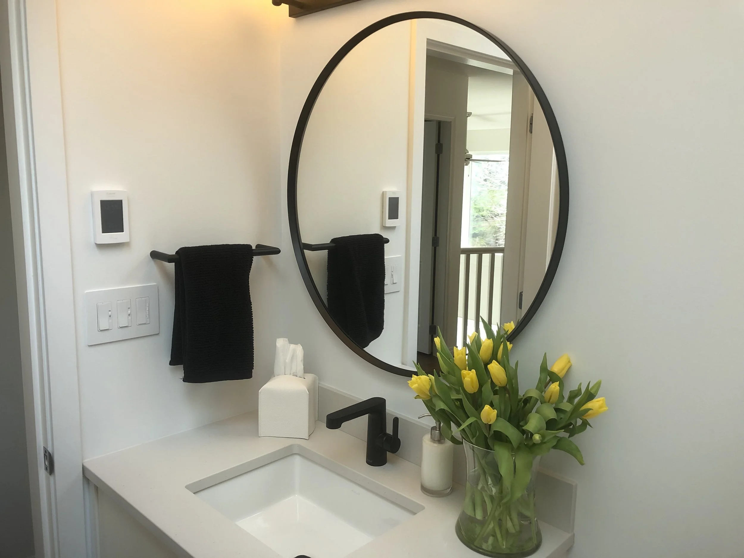 A bathroom sink area with a large oval mirror, a white countertop, and a vase of yellow tulips. Two black towels hang on a towel bar, and a tissue box is on the counter. There are wall-mounted control panels and a window with a balcony outside.