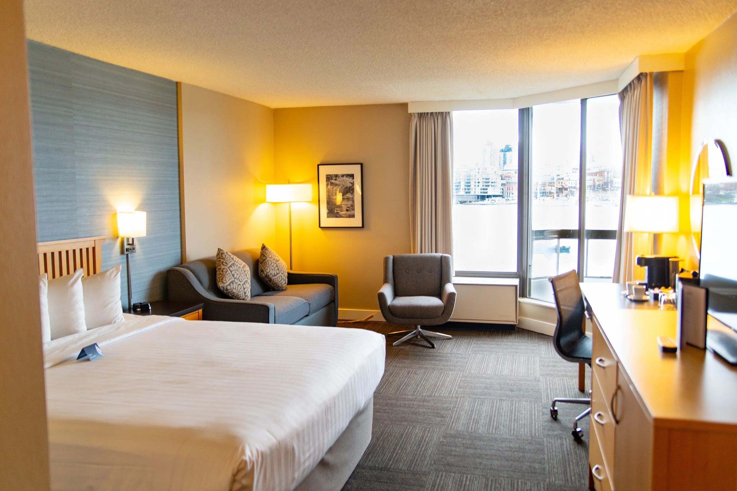 A hotel room with a large bed, a sitting area with a sofa and an armchair, a desk with a chair, and a window showing a city view with buildings and a marina.