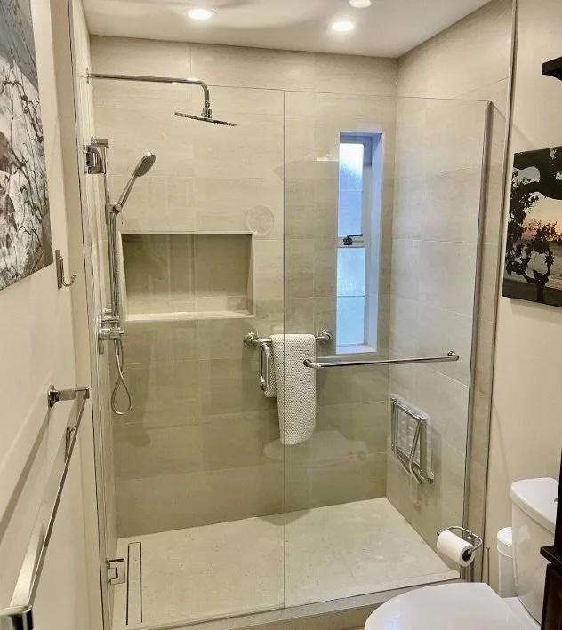 A modern walk-in shower with glass doors, a handheld showerhead, a built-in shelf, a small window, and safety grab bars in a bathroom.