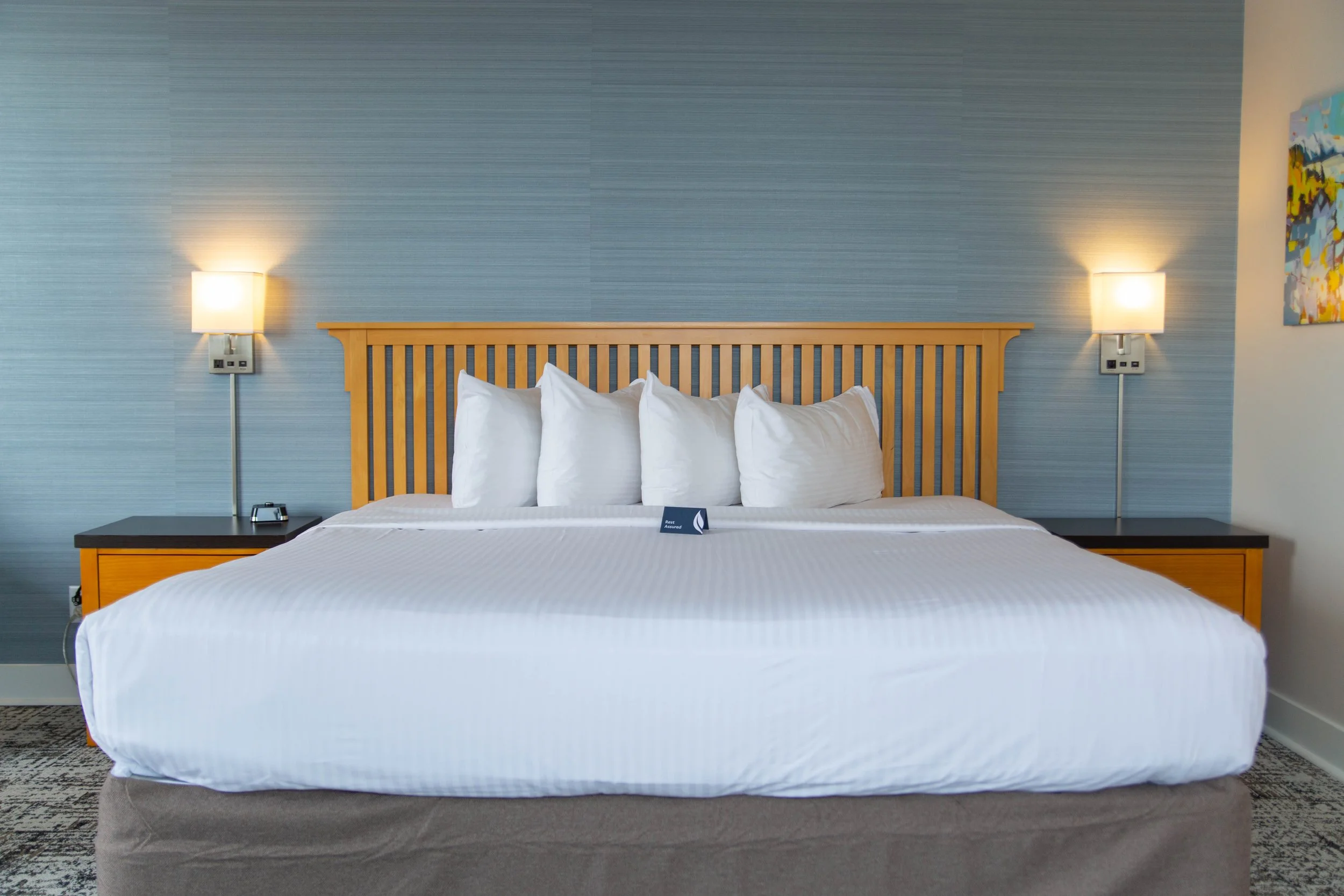 A neatly made hotel bed with white linens and multiple pillows against a wooden headboard. There are two wall-mounted lamps on either side of the bed, and a side table with an alarm clock and phone. The wall behind the bed has a textured blue-grey wa