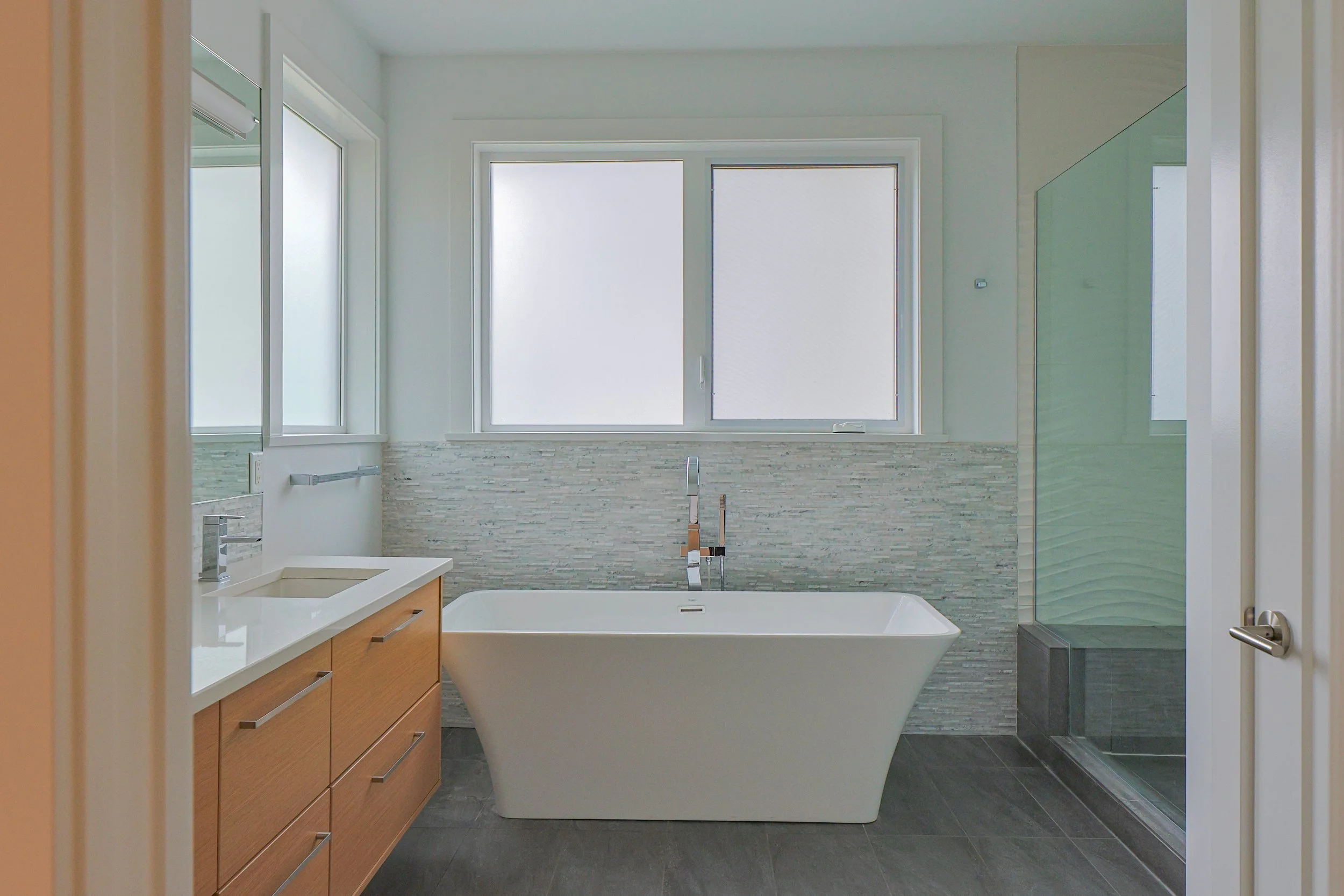 Modern bathroom with a large bathtub, wooden vanity with drawers, multiple windows, a walk-in shower with glass door, and gray tile flooring.