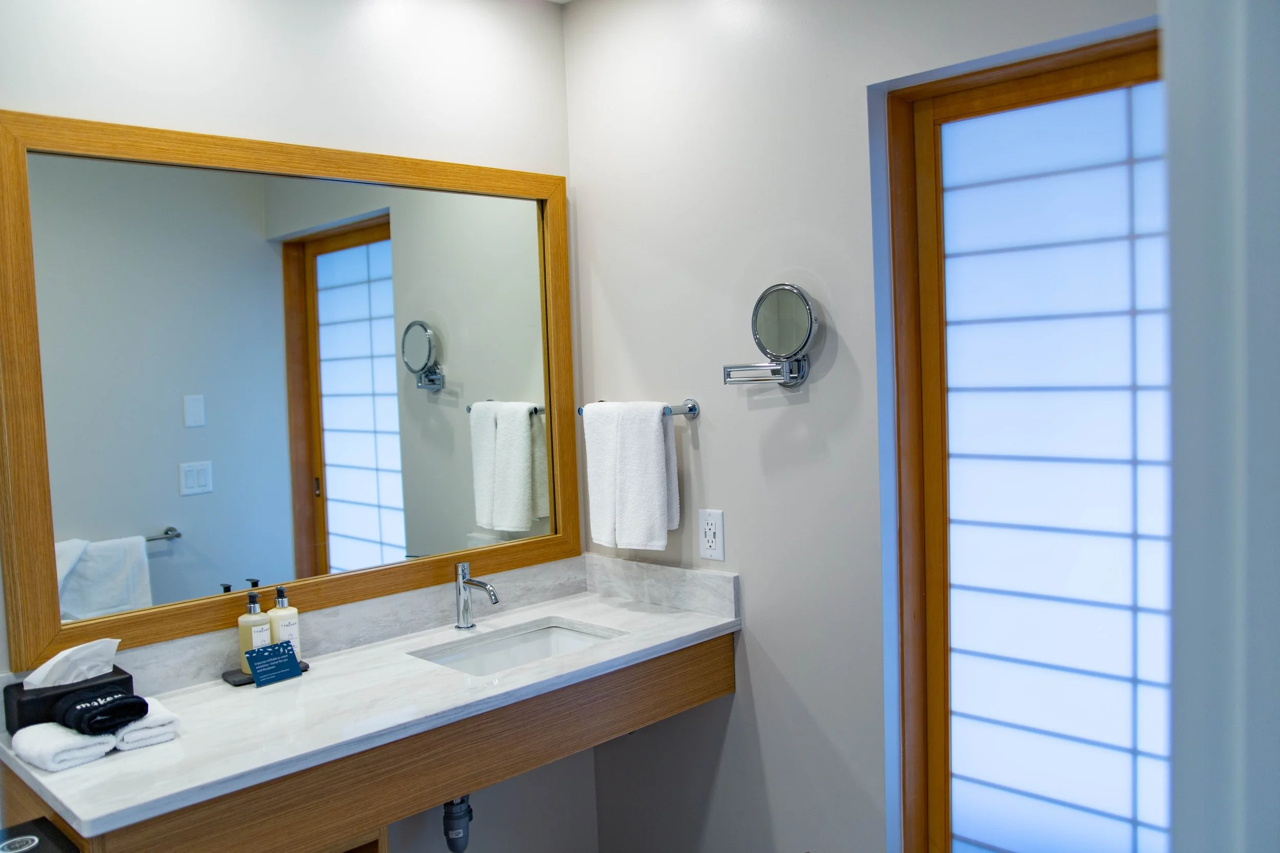 A bathroom with a large mirror above a sink, a window with frosted glass, and a countertop with toiletries, towels, and a tissue box.