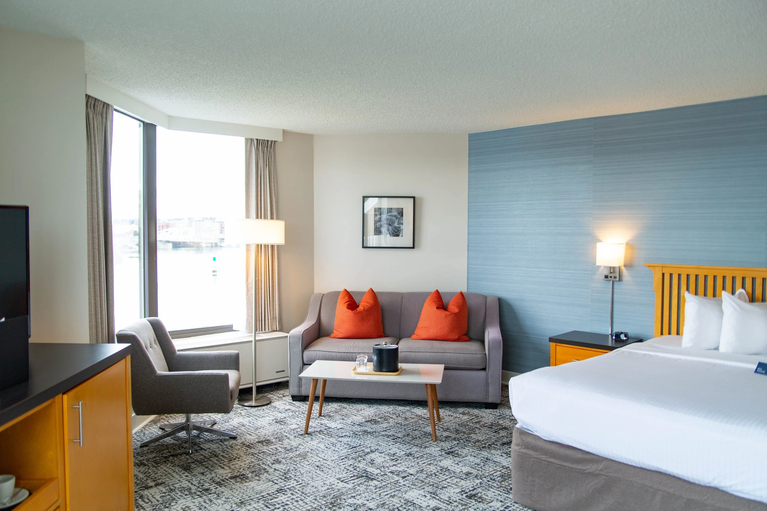 A hotel room with a bed, a gray sofa with orange pillows, a small coffee table, a floor lamp, a framed black and white photo, and a large window with curtains, a nightstand with a lamp, and a view of city buildings outside.