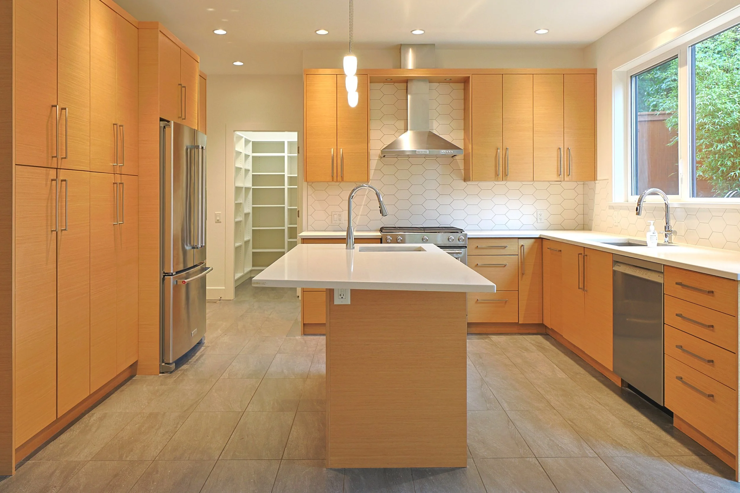 Modern kitchen with light wood cabinets, stainless steel appliances, a center island with a sink, and a large window overlooking trees.
