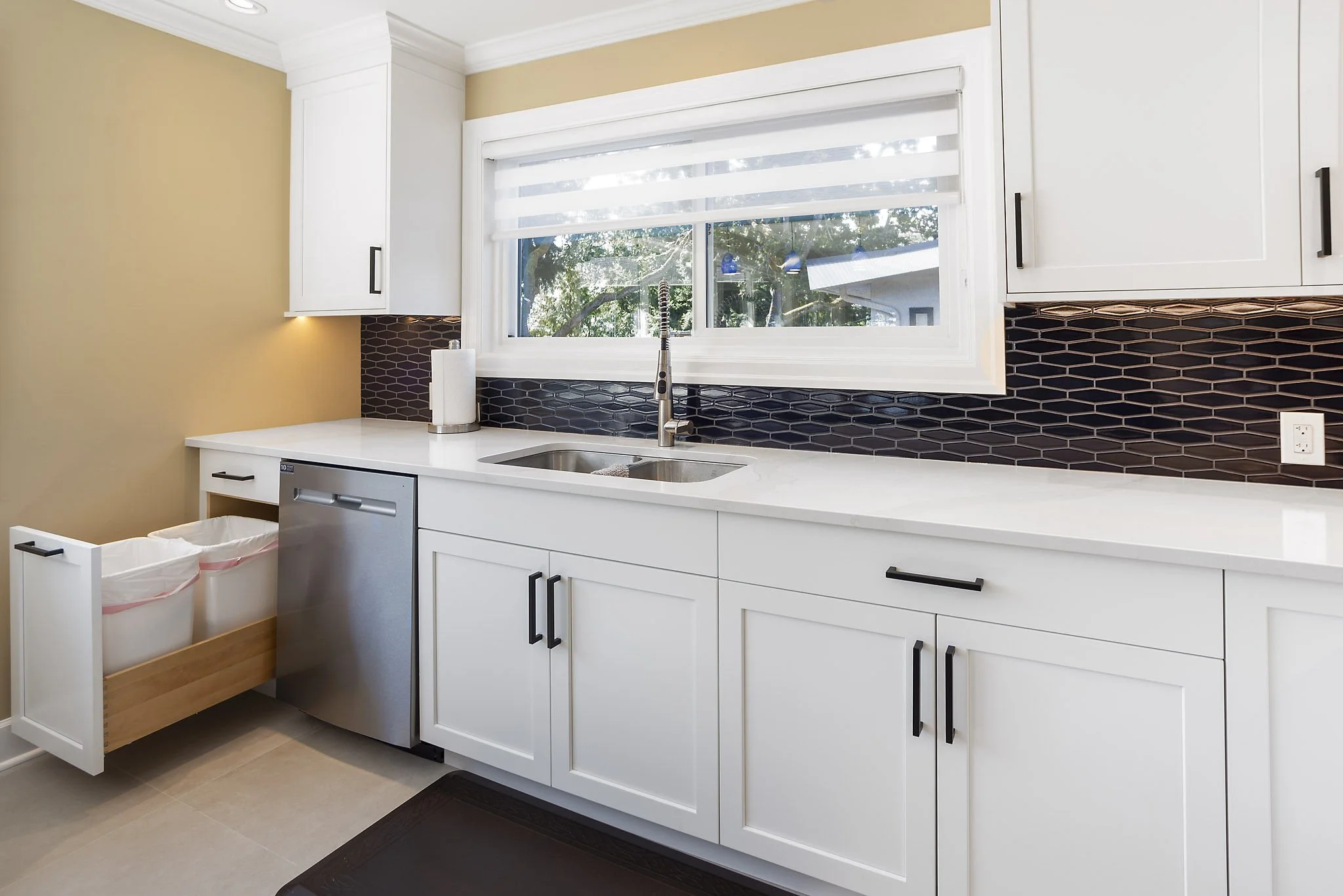 Kitchen with white cabinets, black handles, a window with blinds above a white countertop, a black backsplash, a stainless steel sink, a paper towel holder, and two trash bins.