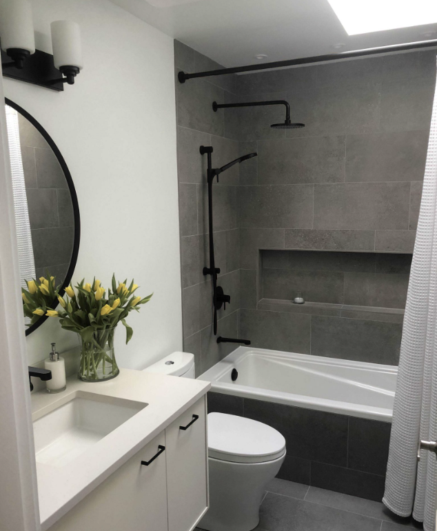 Modern bathroom with gray tiled shower and bathtub, white vanity with black hardware, oval mirror, white toilet, and a vase of yellow tulips on the vanity.