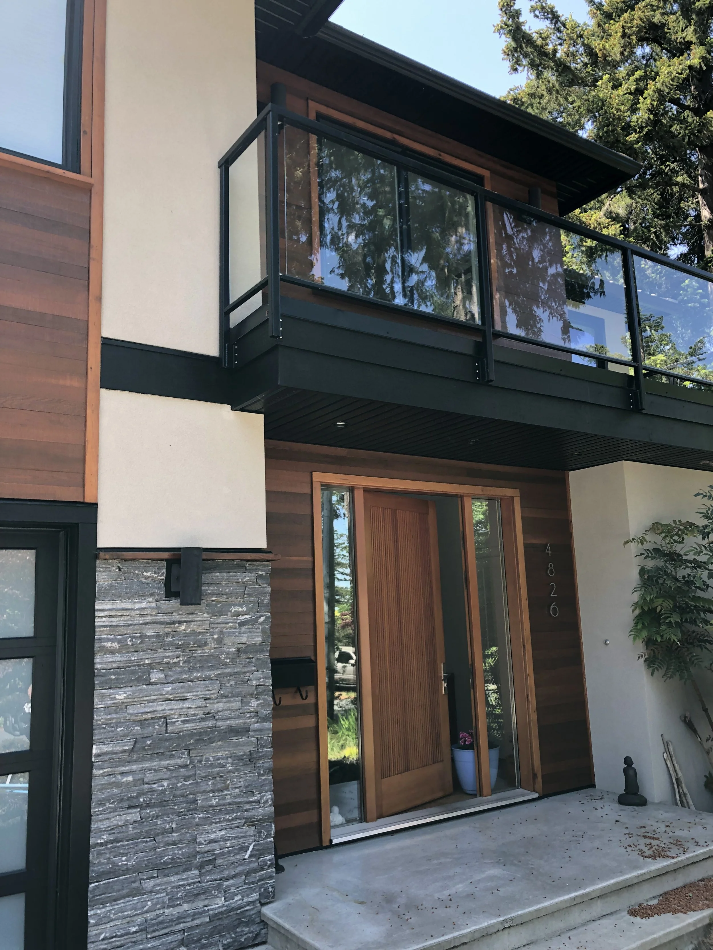 Close-up of a modern house entrance with a wooden door, glass panels, a small balcony with glass railing, and exterior wall with stone and wood siding.