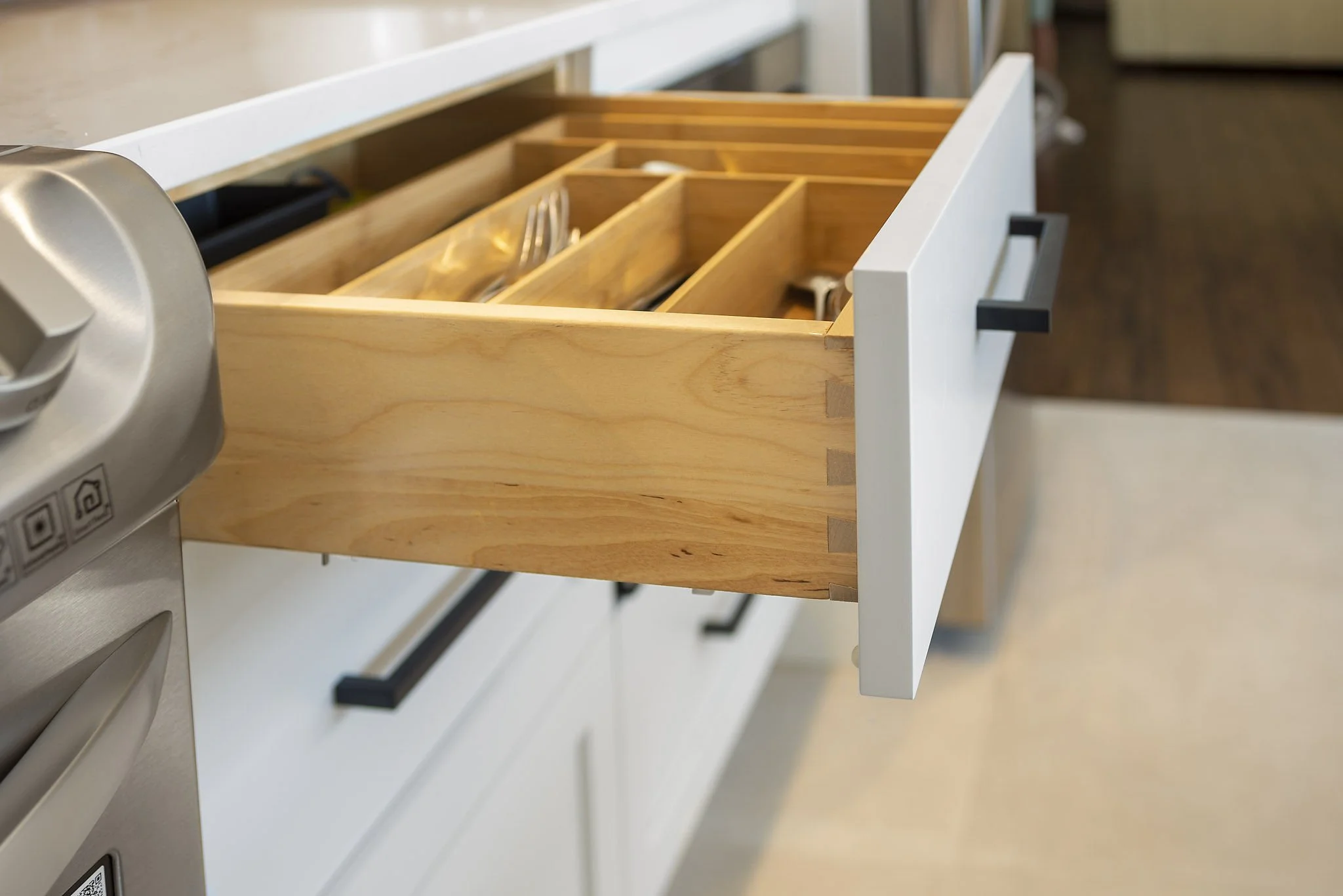 Open kitchen drawer with wooden dividers and utensils inside, adjacent to a white countertop and kitchen appliances.