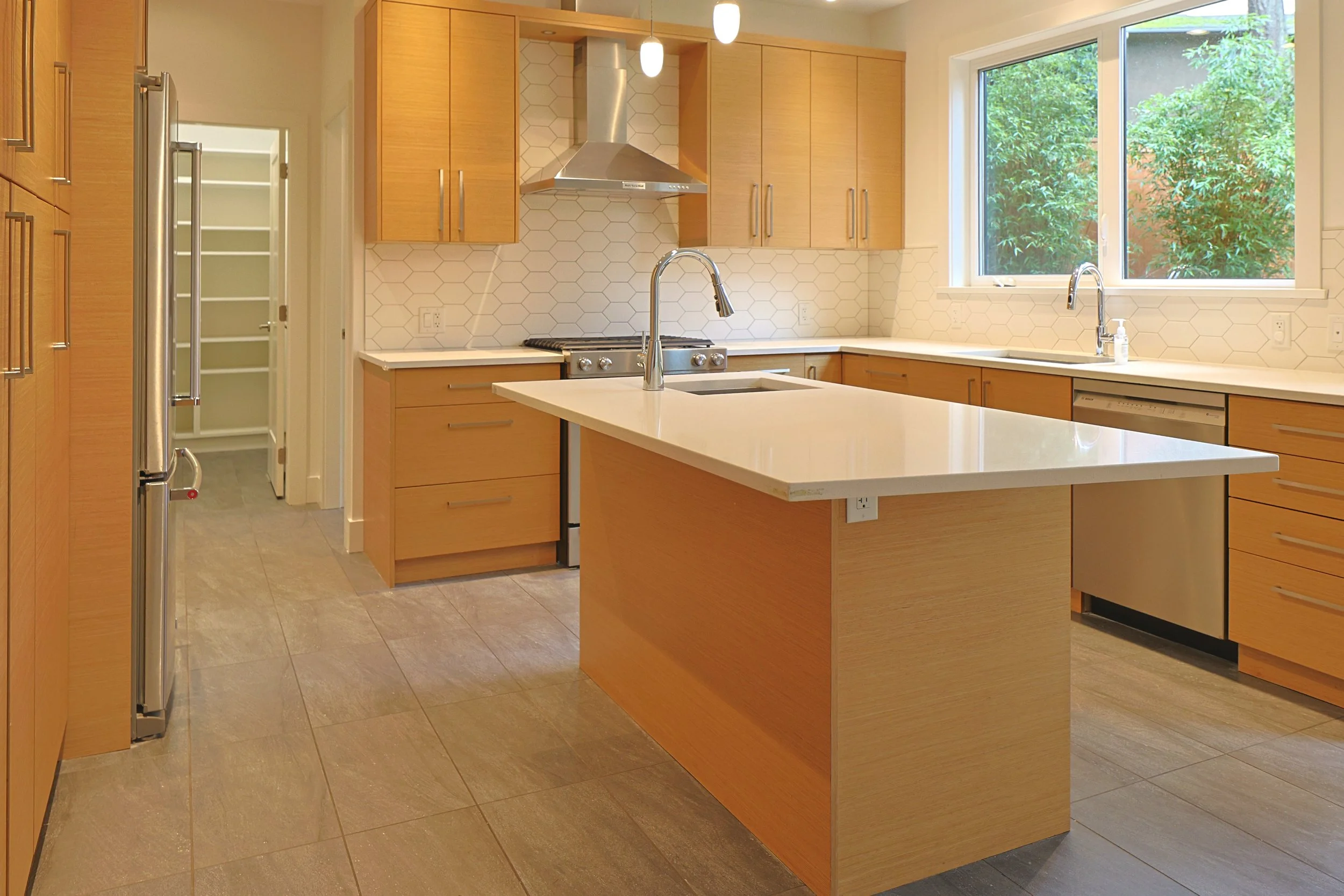 Modern kitchen with wooden cabinets, white countertops, a kitchen island with a sink and dishwasher, large window showing greenery outside, stainless steel range hood and appliances, and a pantry with open door.
