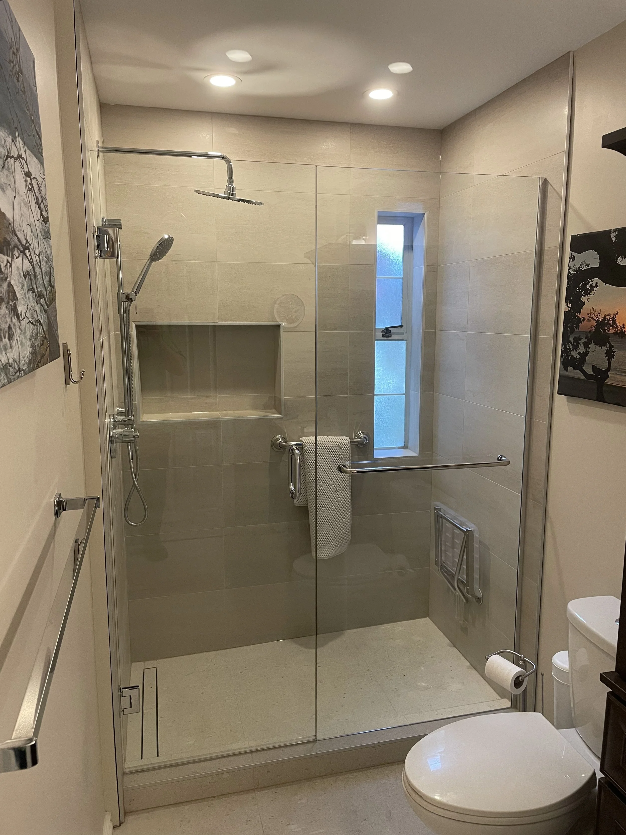 Modern bathroom with shower area enclosed by a glass door, a small window, and a toilet. Towel hanging on a grab bar, artwork on the walls, and a toilet paper holder.