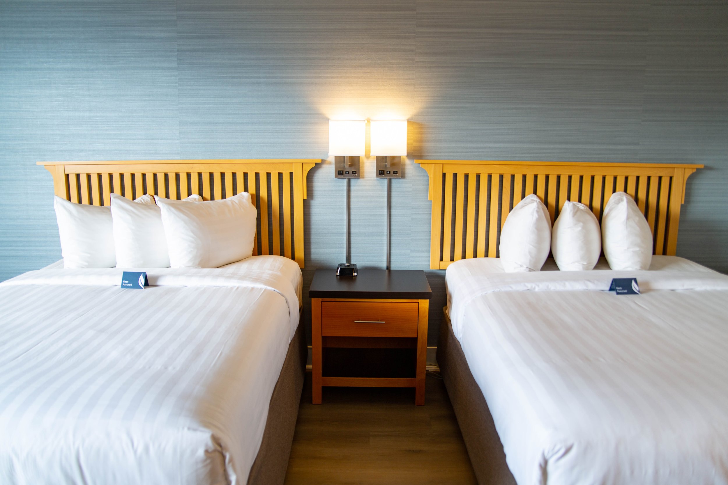 Two neatly made hotel beds with white linens and pillows, separated by a wooden nightstand holding a black lamp. The room has blue textured wallpaper and wall-mounted lamps above each bed.