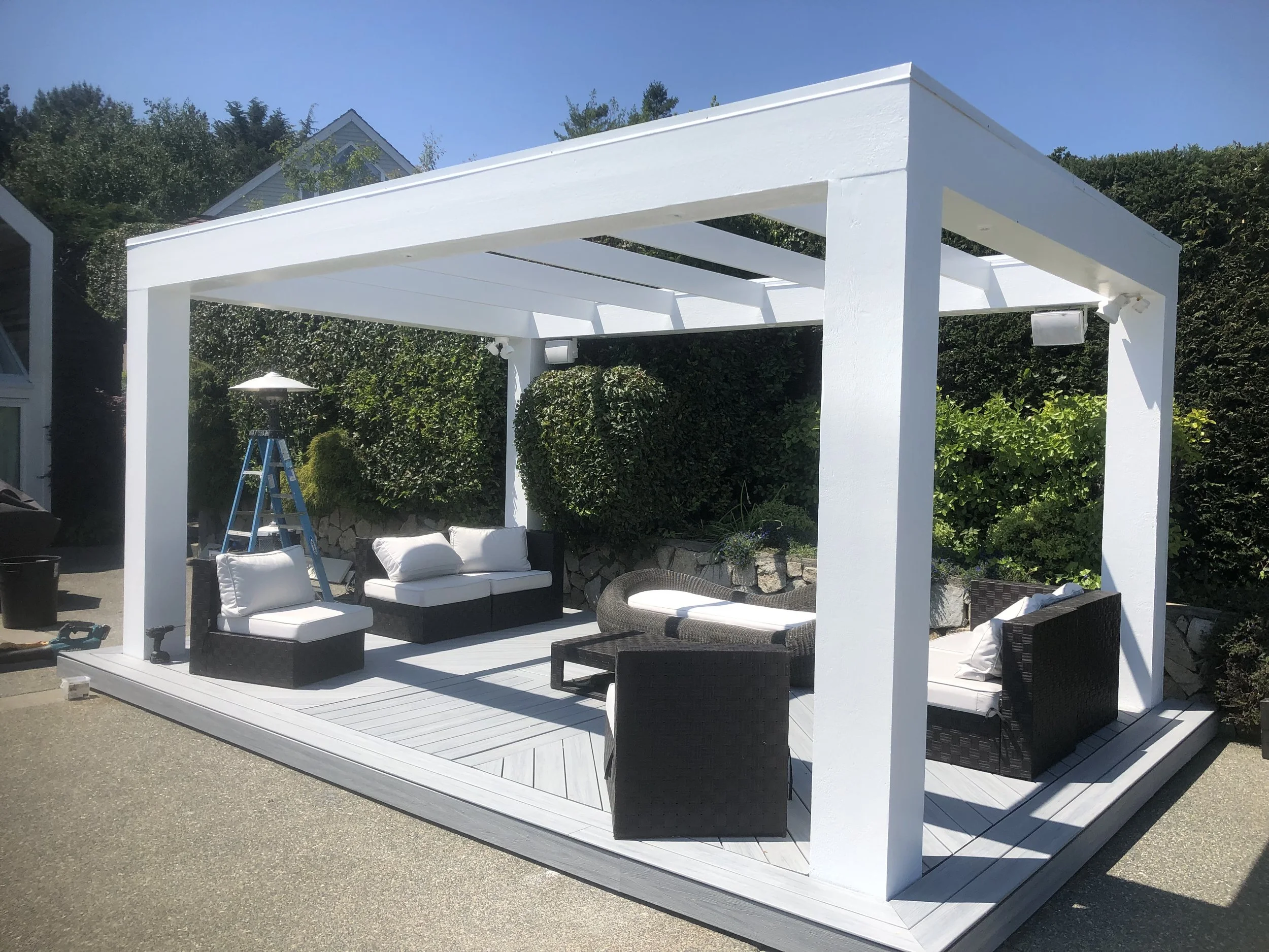 A white wooden pergola on a deck, with outdoor seating including white cushioned furniture and a wicker chair, surrounded by greenery and blue sky.
