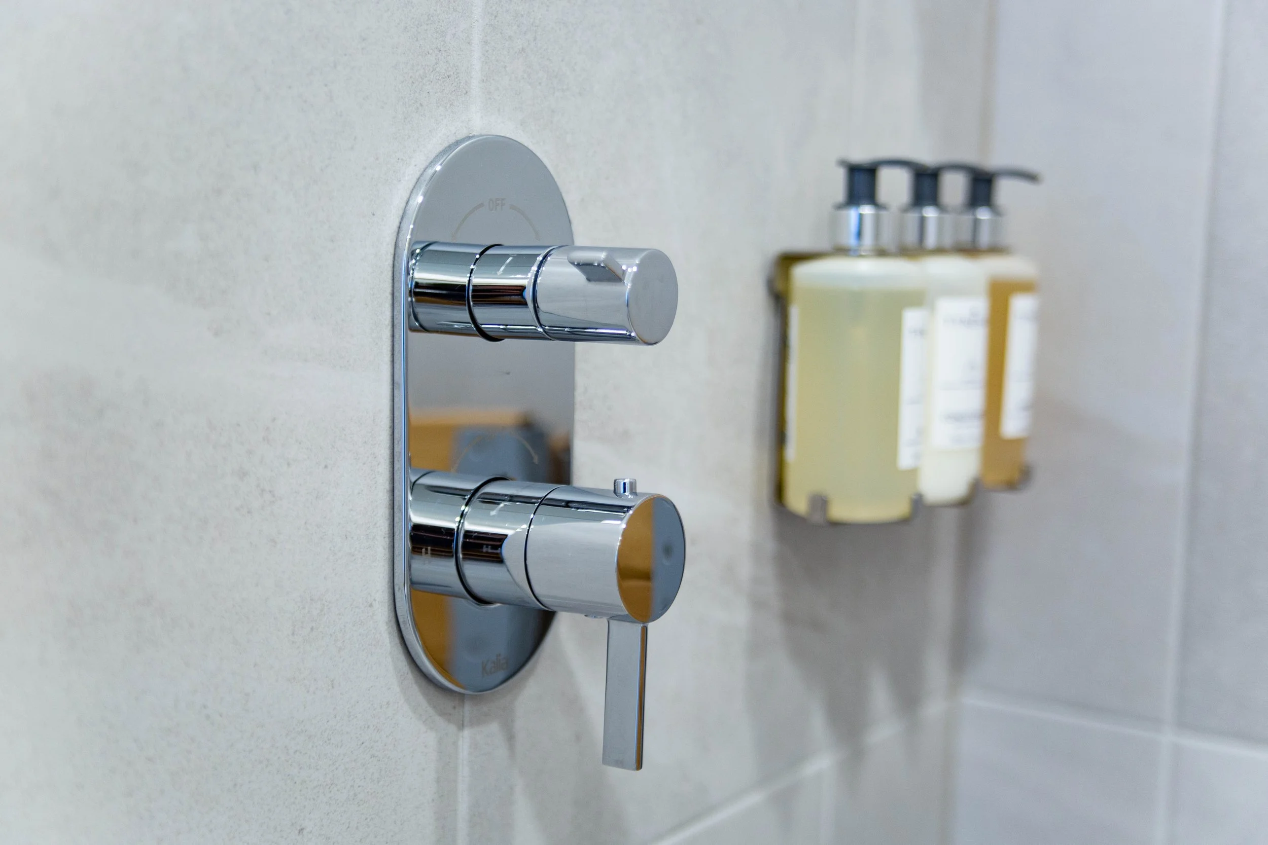 Close-up of a shiny chrome faucet with separate handles for hot and cold water mounted on a light-colored bathroom wall with bottles of toiletries in the background.