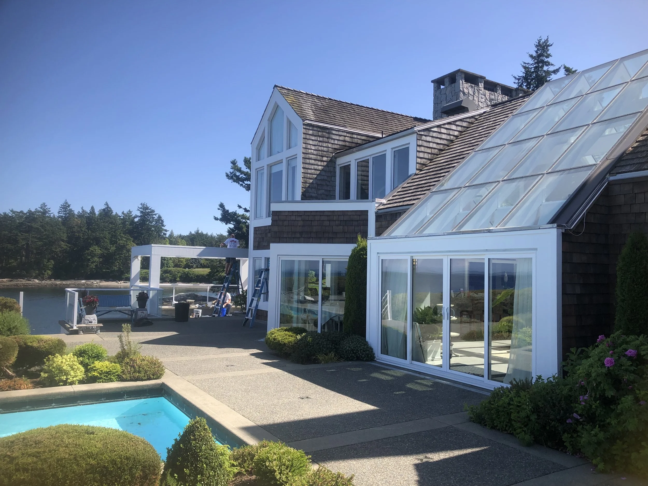 A house with a sloped shingle roof and large windows features a glass-enclosed sunroom. The outdoor area includes a swimming pool, patio furniture, and lush landscaping with bushes and flowers, set against a backdrop of trees and a clear blue sky.