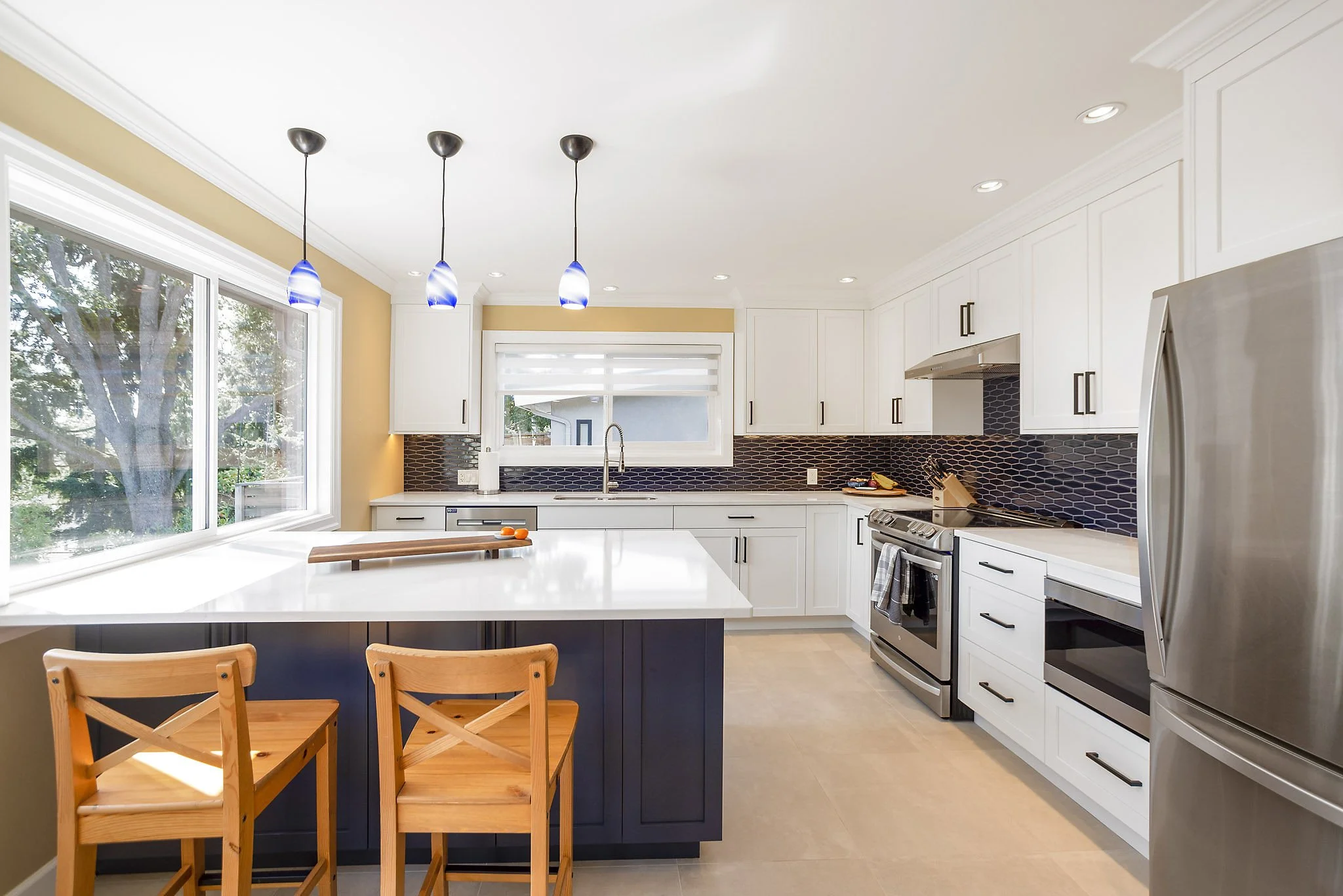 Modern white kitchen with black backsplash, large window, and three pendant lights over a white island with wooden chairs.
