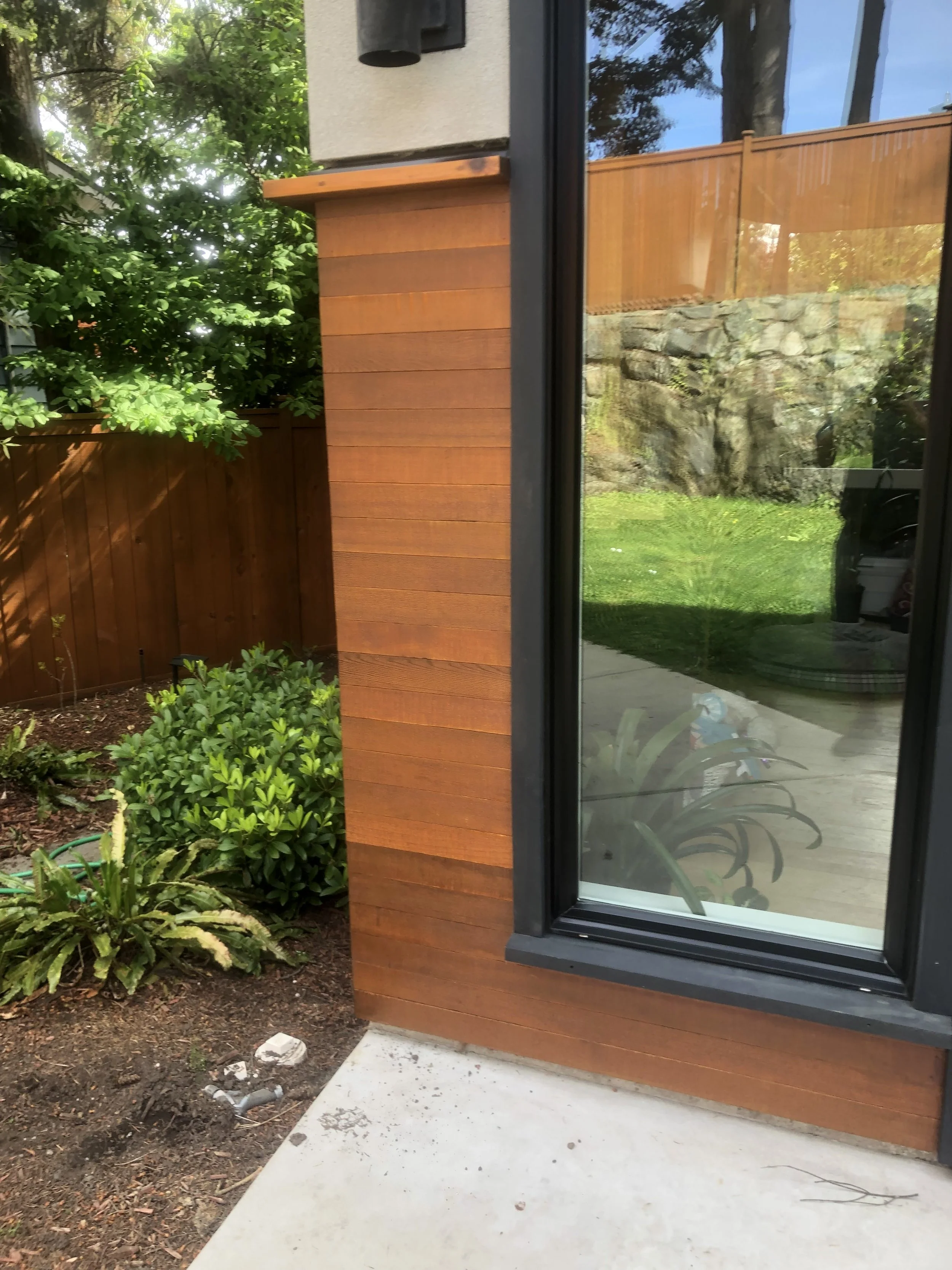 View of a backyard with a mix of green plants, a wooden fence, a stone retaining wall, and part of a modern house with large glass windows. The photo is taken from inside the house looking out through a window.