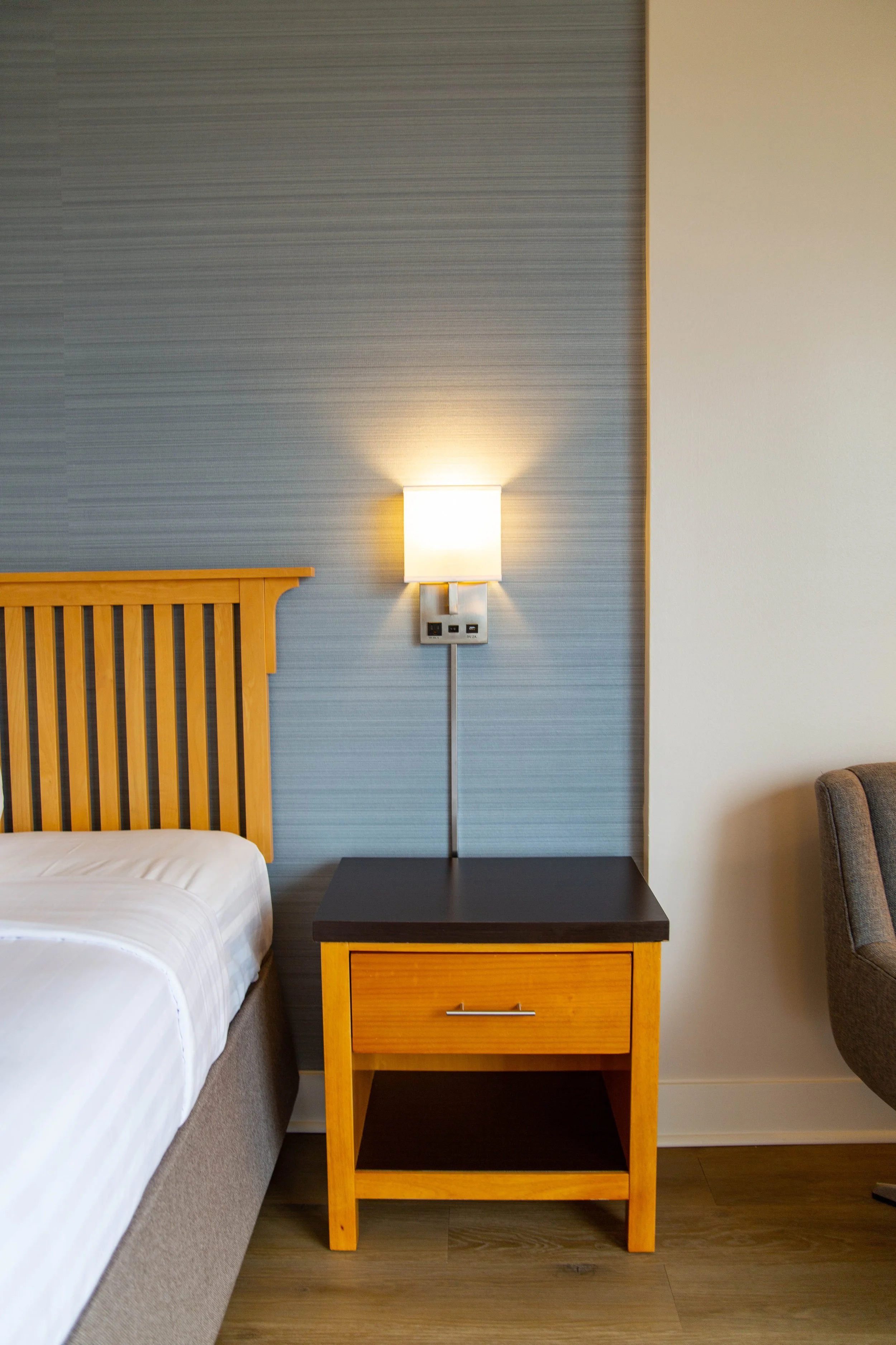 A hotel room nightstand with a wooden frame, a black top surface, and a lower shelf, placed next to a bed with a wooden headboard and white bedding. A wall-mounted light fixture is above the nightstand, and the wall behind has a textured blue-gray an