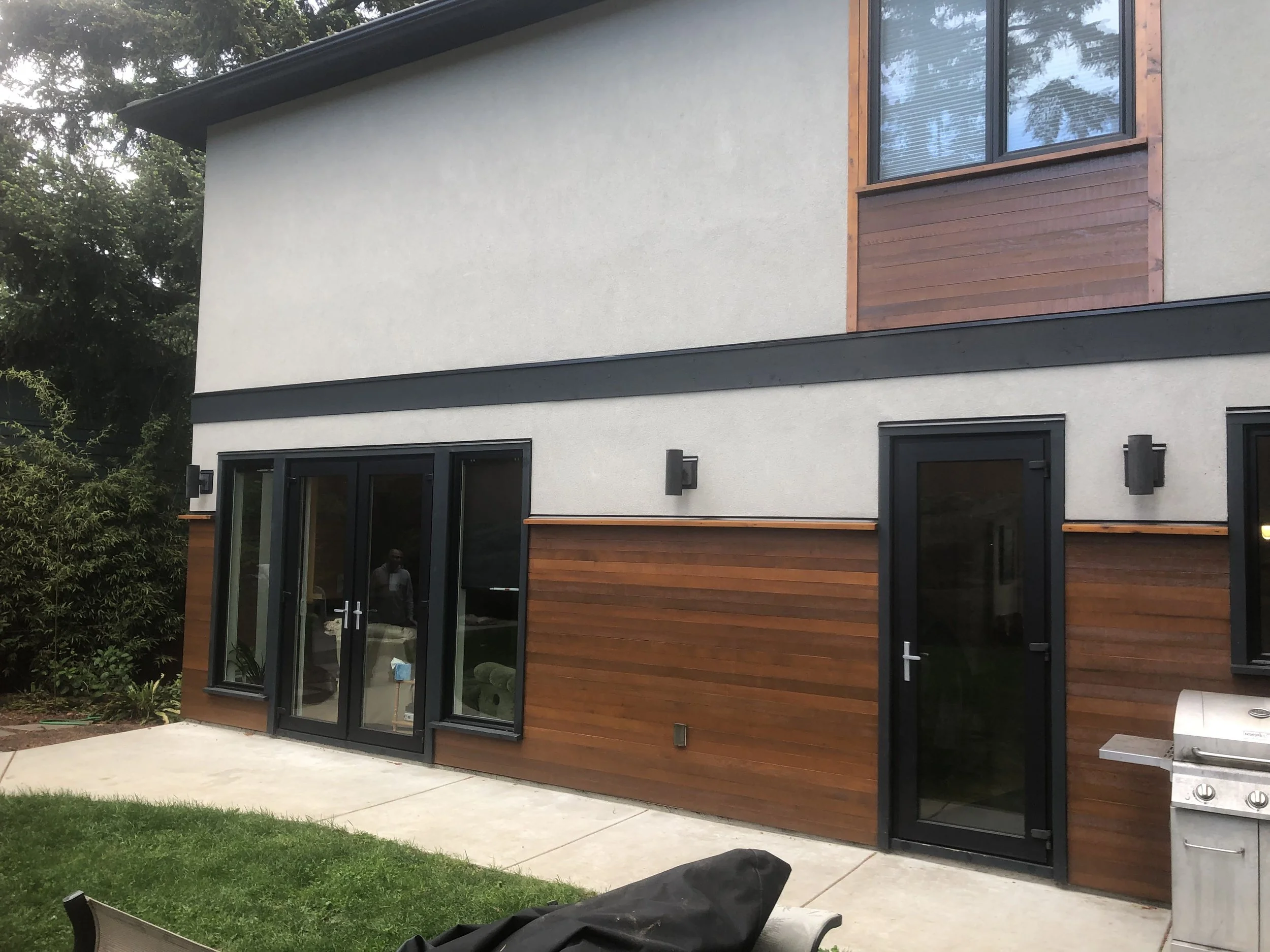 A modern house with a combination of white stucco and wooden siding, large glass doors, and windows, with outdoor lighting fixtures attached to the wall.