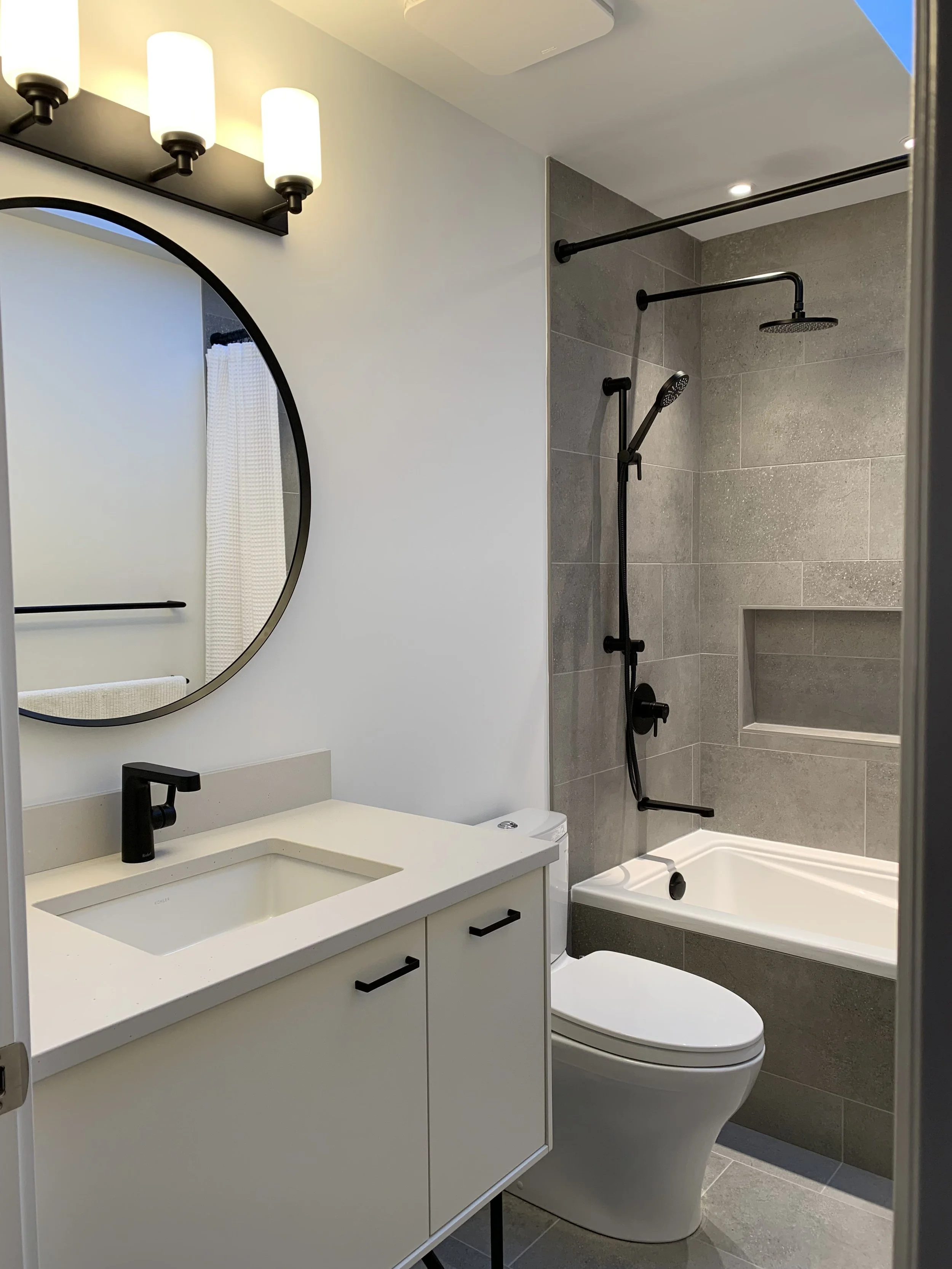 Modern bathroom featuring a vanity with a white countertop, black faucet, and a round mirror, alongside a shower area with gray tiles, a ceiling-mounted showerhead, a handheld shower wand, and a built-in shower niche.