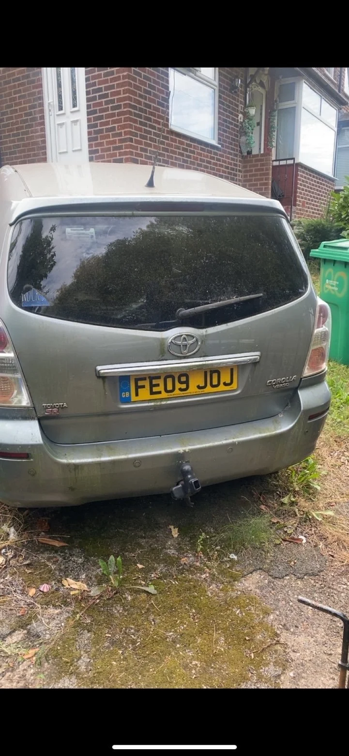 The rear of a silver Toyota Corolla Verso with a UK license plate FE09 JJJ, parked on a patchy dirt and grass surface in front of a brick house with large windows and a white door.